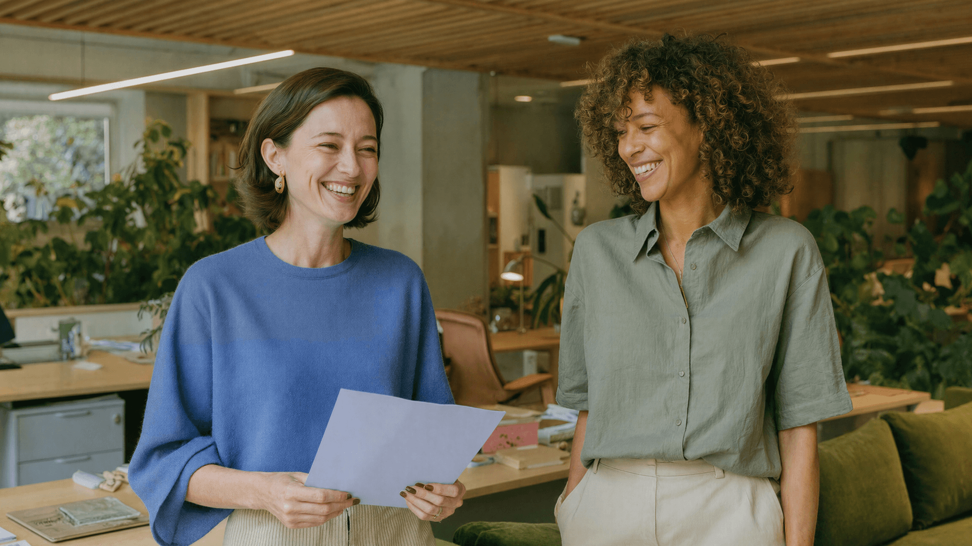 Duas mulheres sorrindo enquanto analisam um documento juntas em um escritório moderno cercado de plantas, representando a comunicação eficiente entre profissionais de clínicas com o suporte da Tivita.