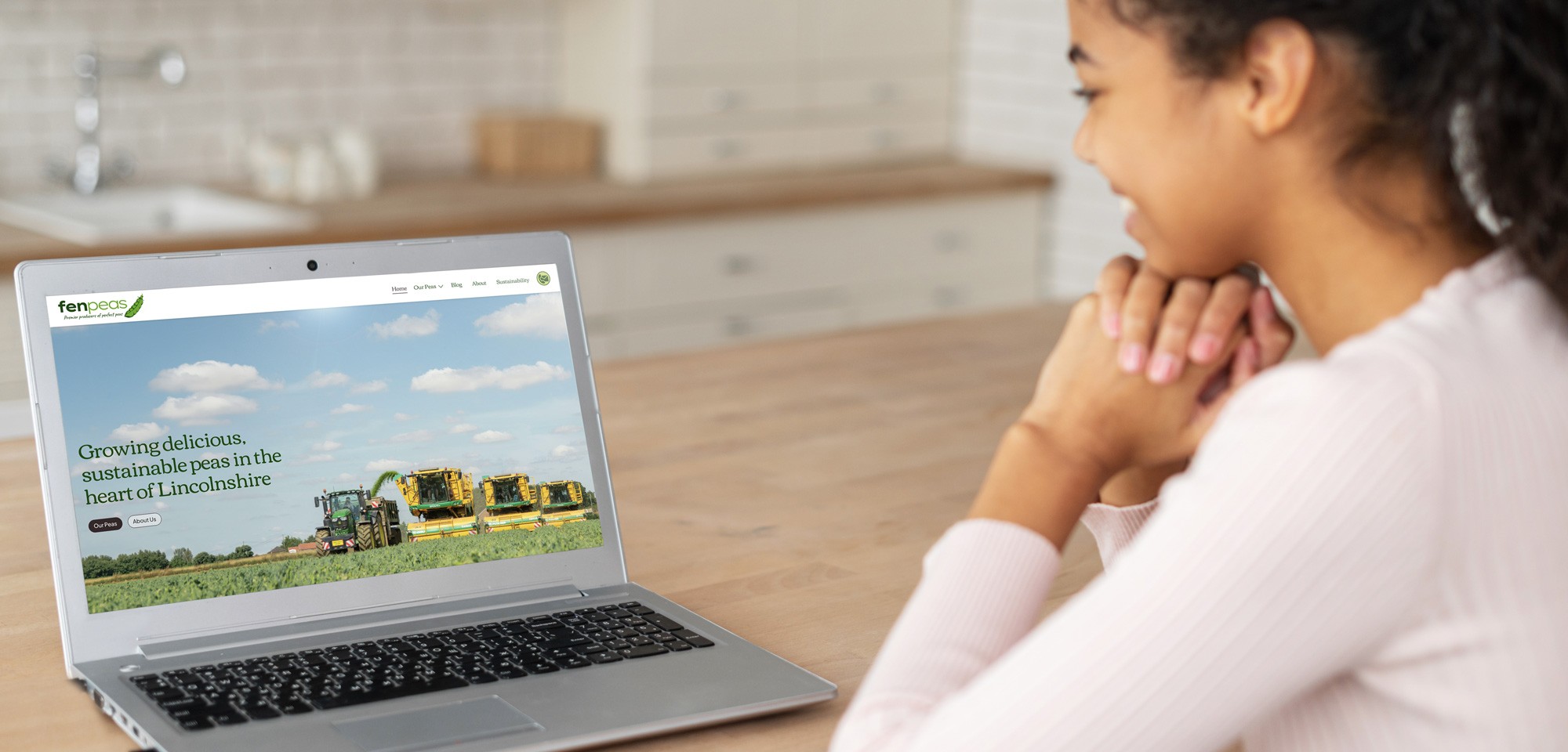 Teenage girl at a kitchen table looking at a laptop showing the Fen Peas homepage, headlined "Growing delicious, sustainable peas in the heart of Lincolnshire" over a photo of pea harvesting machinery in a field.