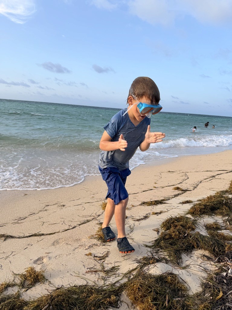 A child wearing a snorkel mask on a sandy beach beside the sea.