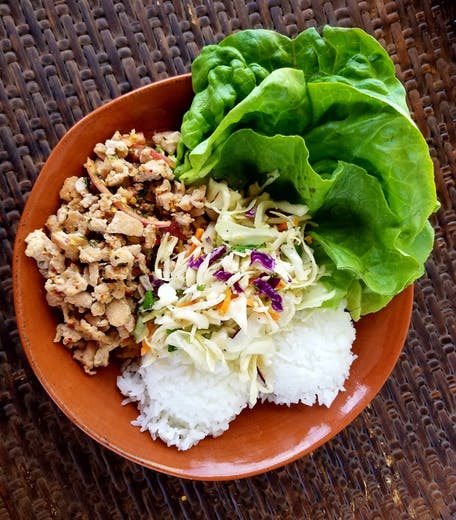 A bowl of seasoned ground chicken larb served with two scoops of white rice, a portion of Asian slaw, and several large, crisp green leaf lettuce leaves.