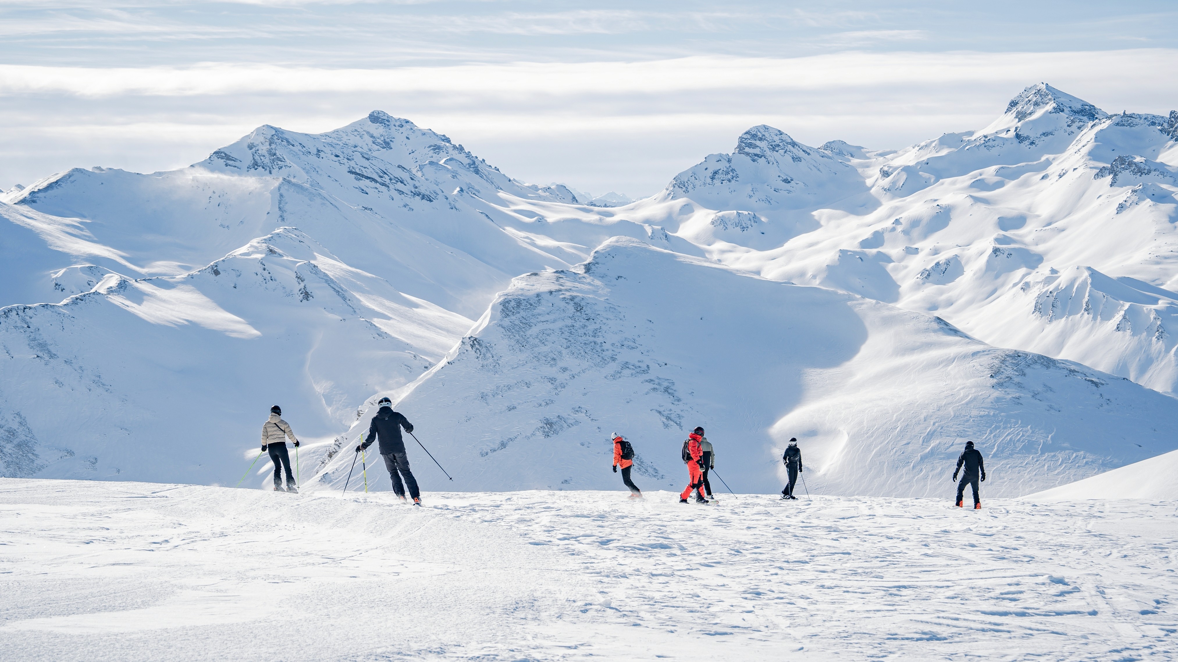 Skiers move across a wide snowy ridge with dramatic, sunlit mountain peaks rising in the background.