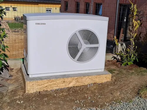 Bílé tepelné čerpadlo s velkým kruhovým větracím otvorem, instalované venku na betonovém podstavci pro energetickou účinnost.