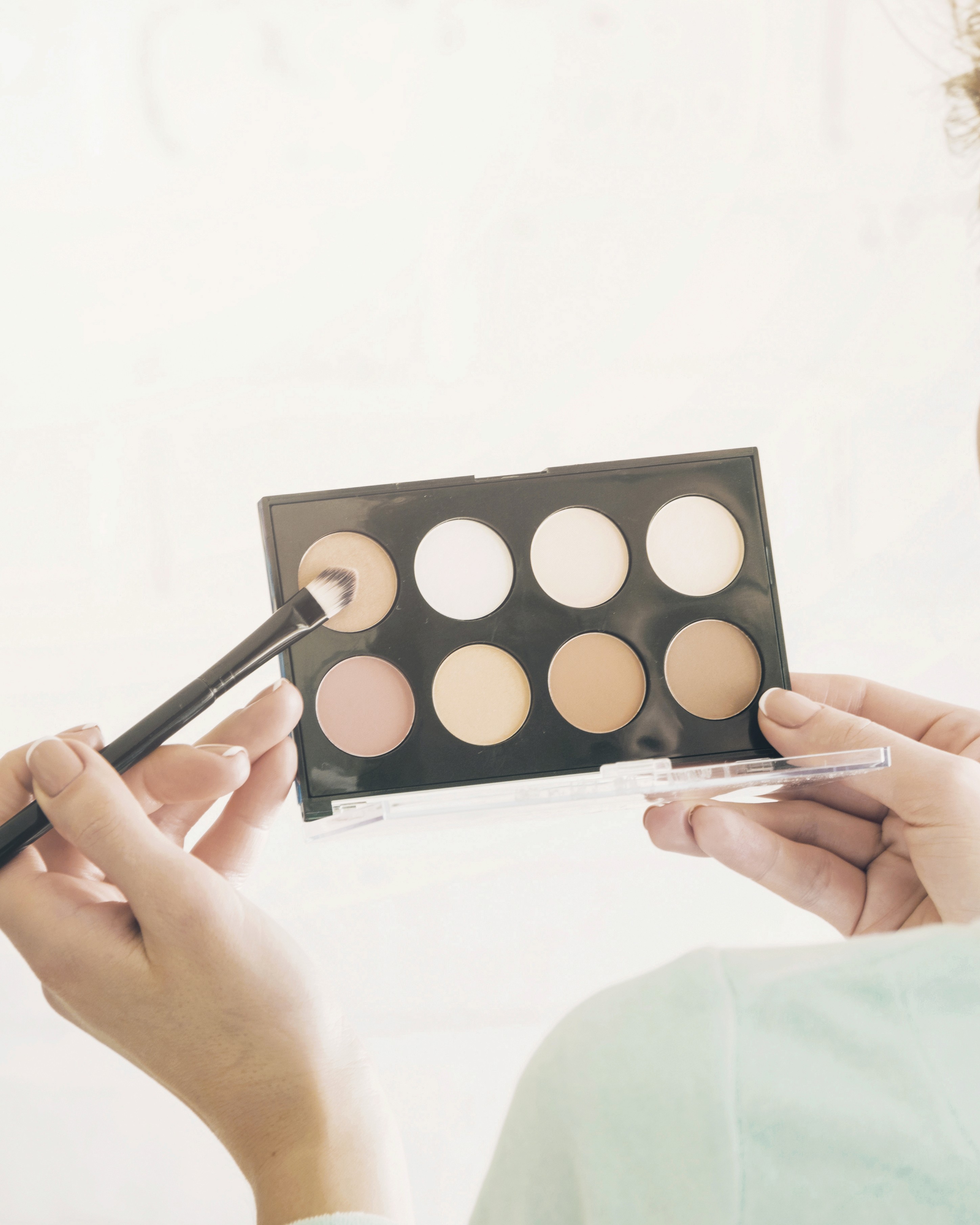 A person applying makeup with a neutral eyeshadow palette and a brush.