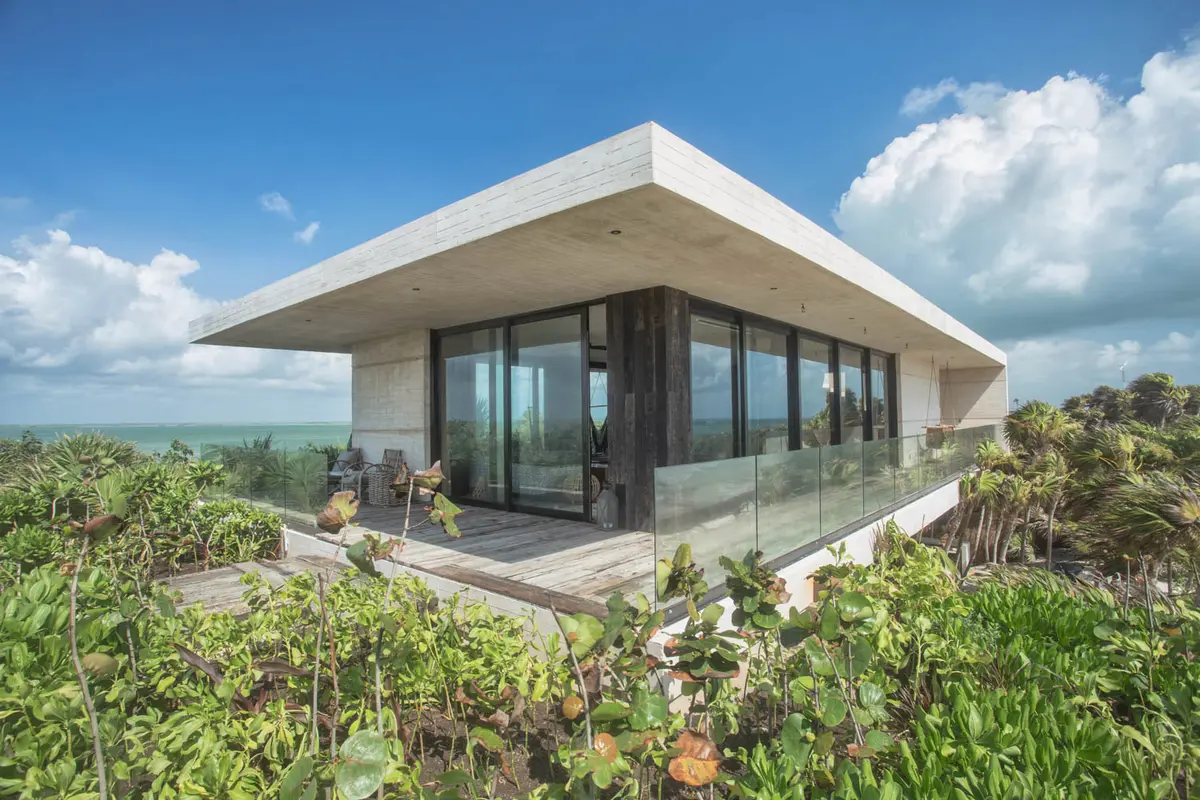 A modern, minimalist house with large glass windows and a spacious deck, surrounded by lush greenery and overlooking the ocean on a sunny day.