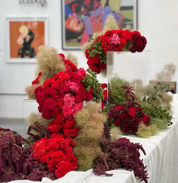Instalación floral contemporánea con flores rojas y fucsias sobre mesas blancas para galerías y artistas que buscan un montaje botánico expresivo y de alto impacto visual.