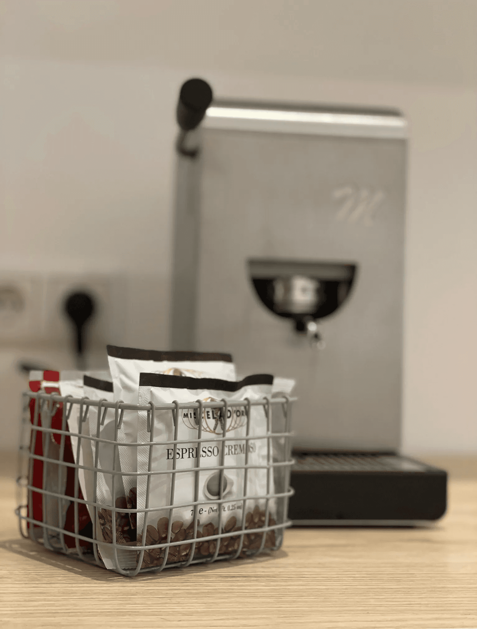 „Metallene Kaffeemaschine auf einer Holztheke mit einem Korb voller Espressobohnen-Päckchen im Vordergrund; minimalistisches und gemütliches Küchenambiente.