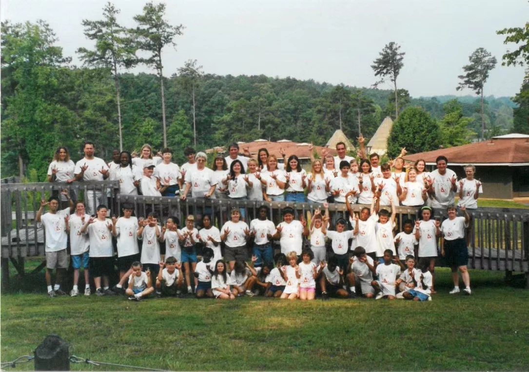 Large group photo of campers and staff at summer camp. Everyone is wearing white Camp Juliena shirts. They are posing on a wooden deck in front of camp buildings.