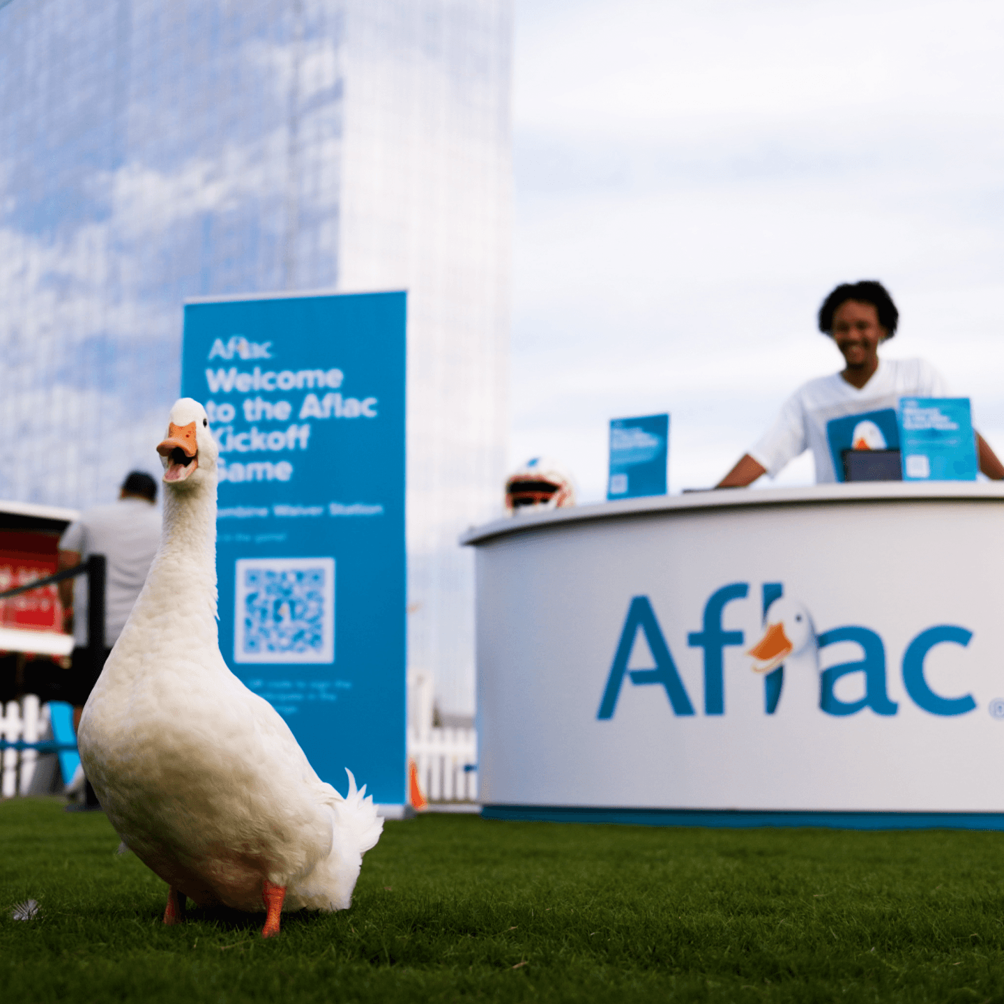 A white duck stands on grass in front of an Aflac booth, with a man smiling behind the counter. Signs with the Aflac logo and event information are in the background, and a tall glass building rises behind them.