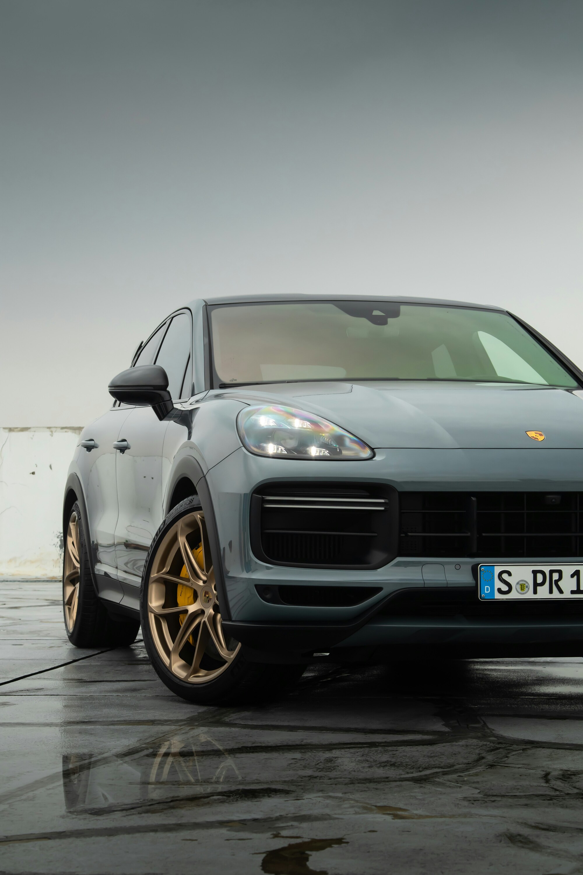 A sleek, modern SUV with a metallic gray finish is parked on a wet concrete surface under an overcast sky, showcasing its distinctive headlights and stylish design, emphasized by large bronze alloy wheels.