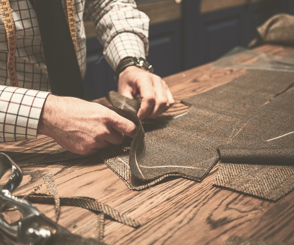 Hands sewing grey fabric on a wooden work table with measuring tape and pattern pieces.