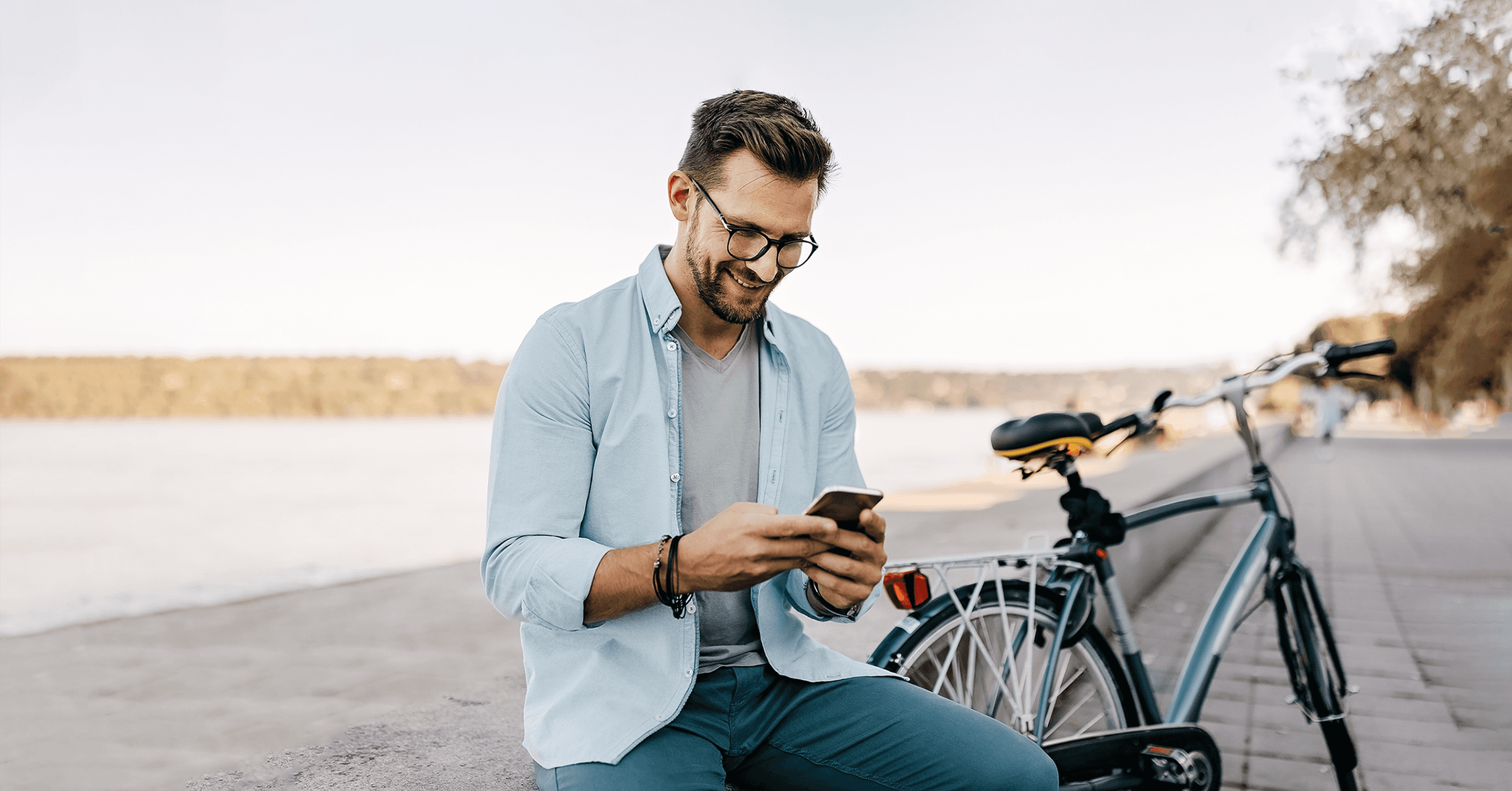 En man sitter ensam vid en sjö och ler mot sin mobil som han håller i handen, och hans cykel står precis bredvid.