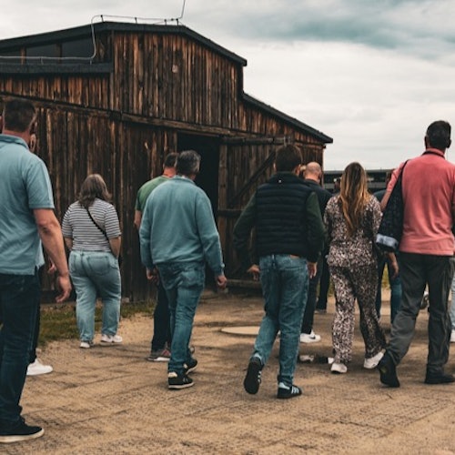 Een groep mensen loopt richting een verweerd houten gebouw op een bewolkte dag.
