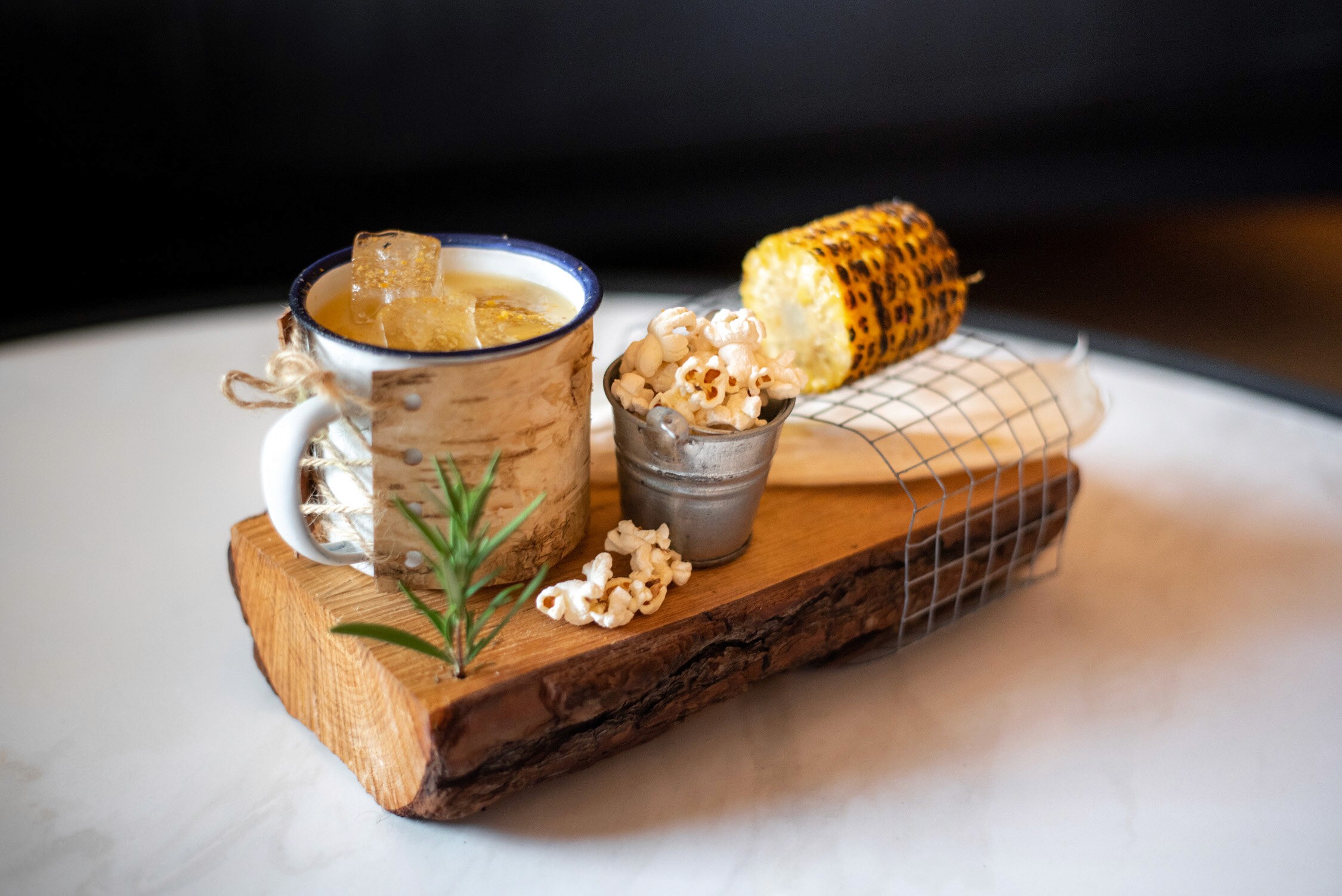 A rustic wooden board holds a mug of hot chocolate with a marshmallow and a small cup of coffee with a piece of toast.