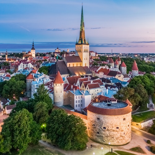 Egy középkori európai város madártávlatból, ahol számos vörös tetős épület, egy nagy templomtorony és környező zöld területek láthatók alkonyatkor.