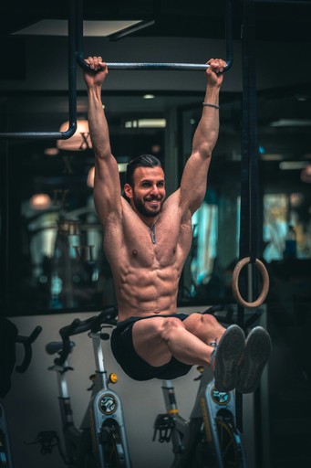 A muscular man smiling while performing a hanging leg raise on gymnastic rings in a bright fitness studio.