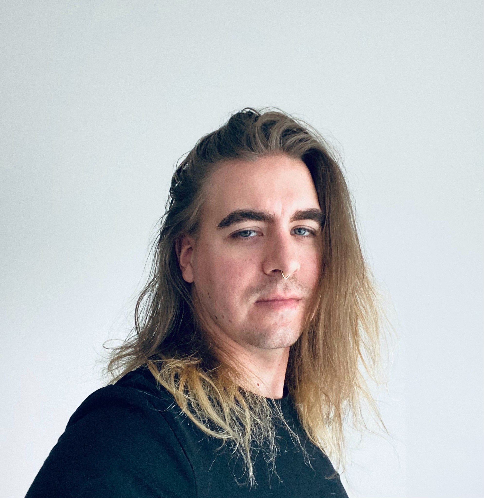 John against a white background. He has long hair open and he is wearing a black t-shirt.