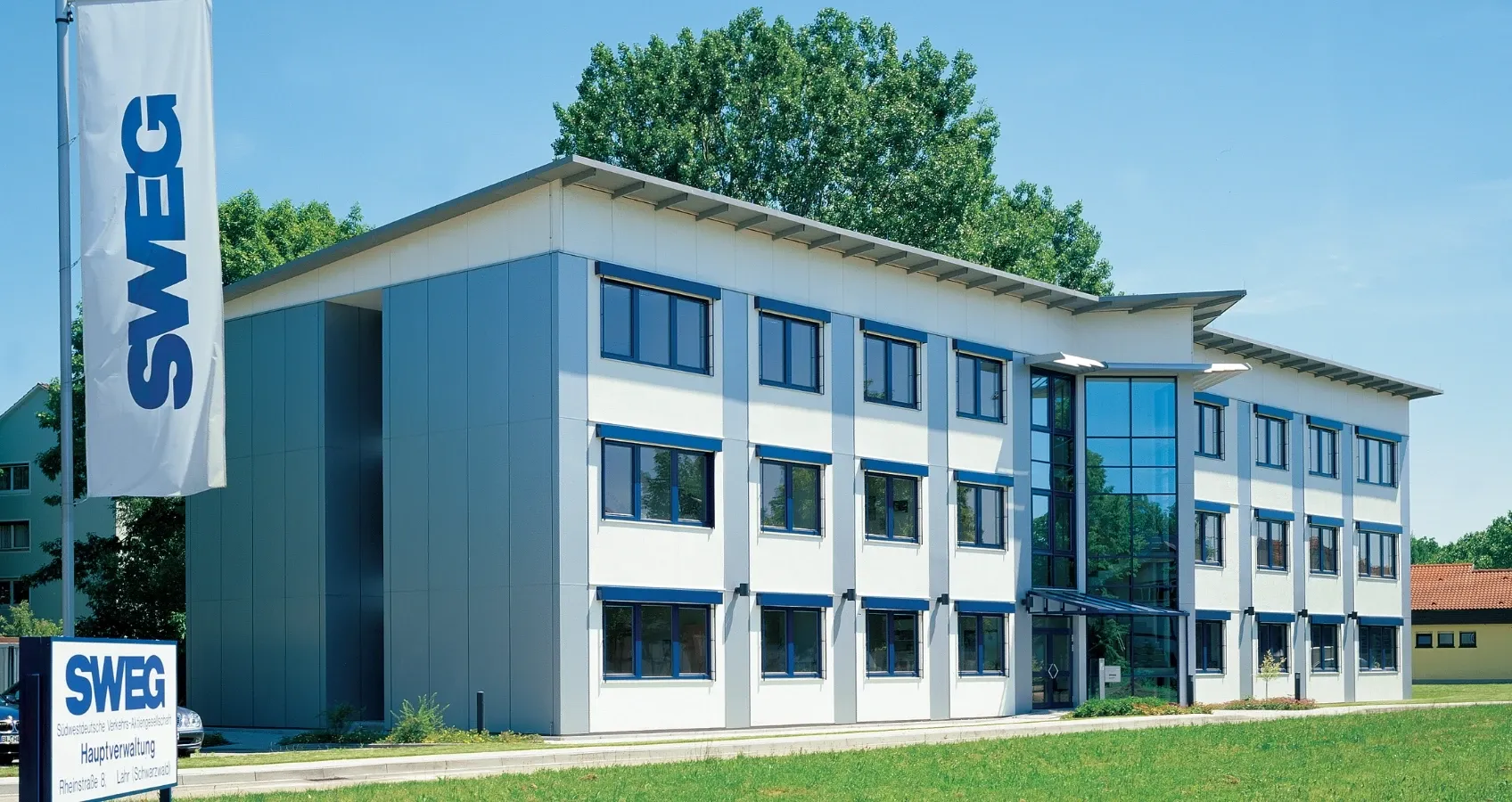 Modernes Verwaltungsgebäude der SWEG mit einem großen Firmenschild und einem Fahnenmast mit blauem Logo.