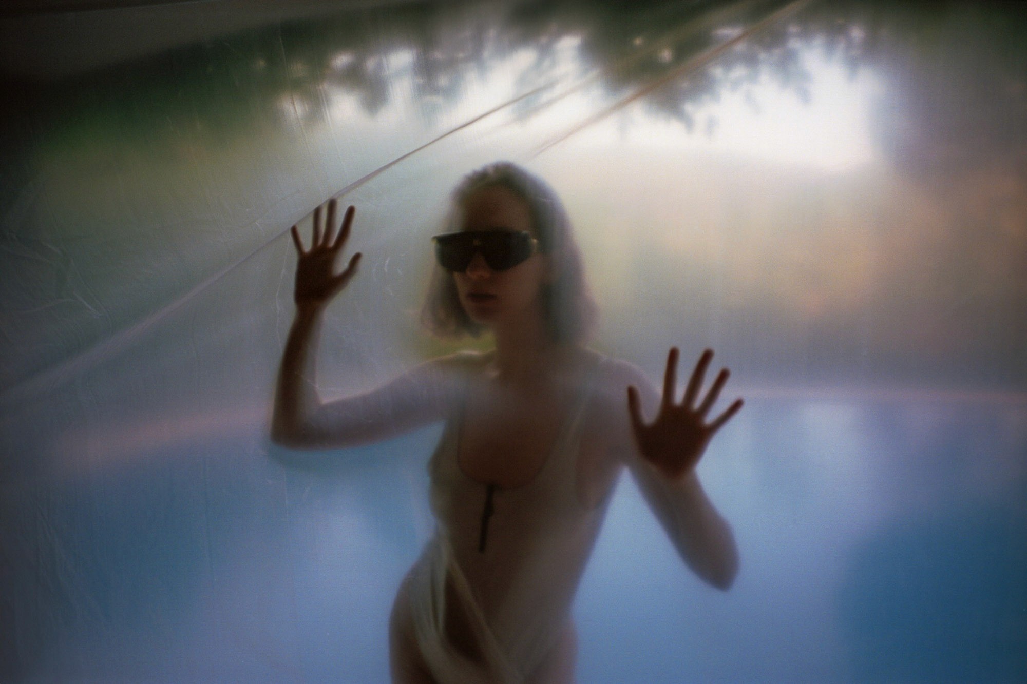 A person stands behind a foggy, translucent surface, wearing sunglasses and a white outfit, with hands pressed against the surface, creating a mysterious and ethereal mood.