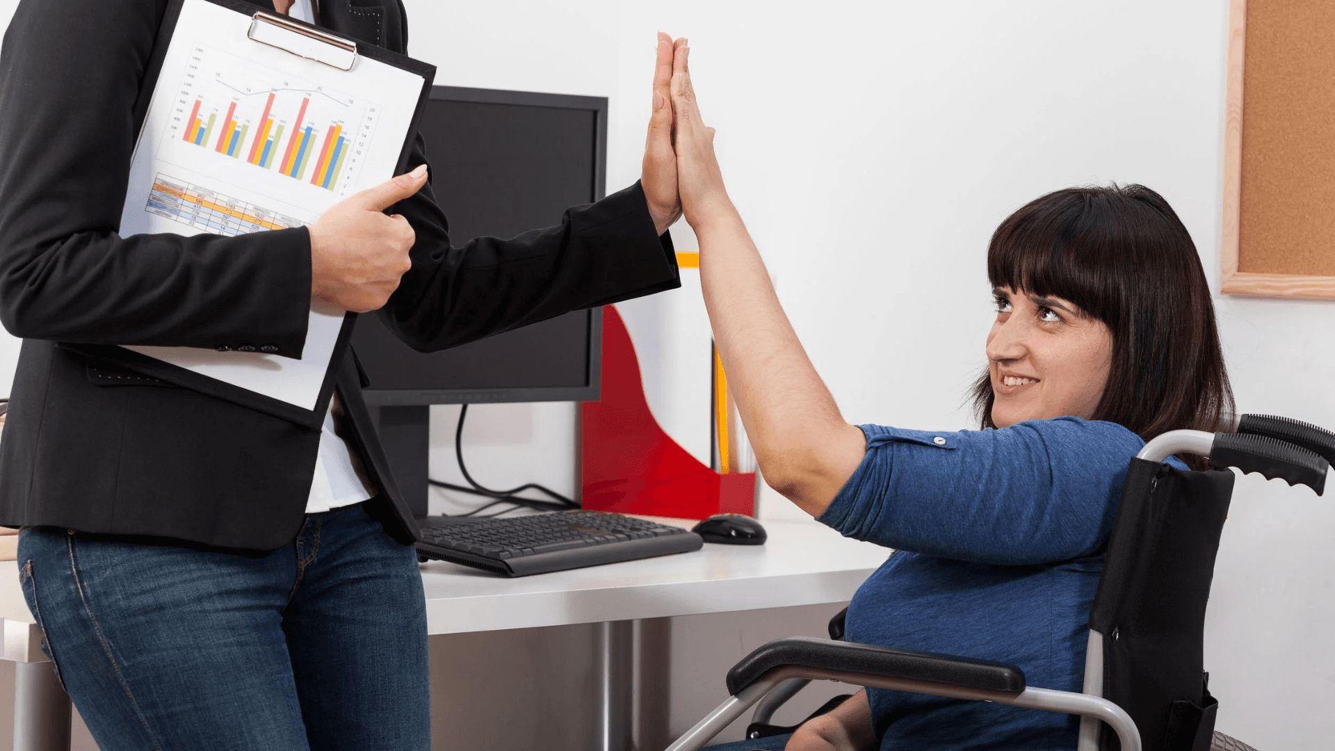 Woman in a wheelchair giving a high-five to a colleague holding a clipboard in an office setting