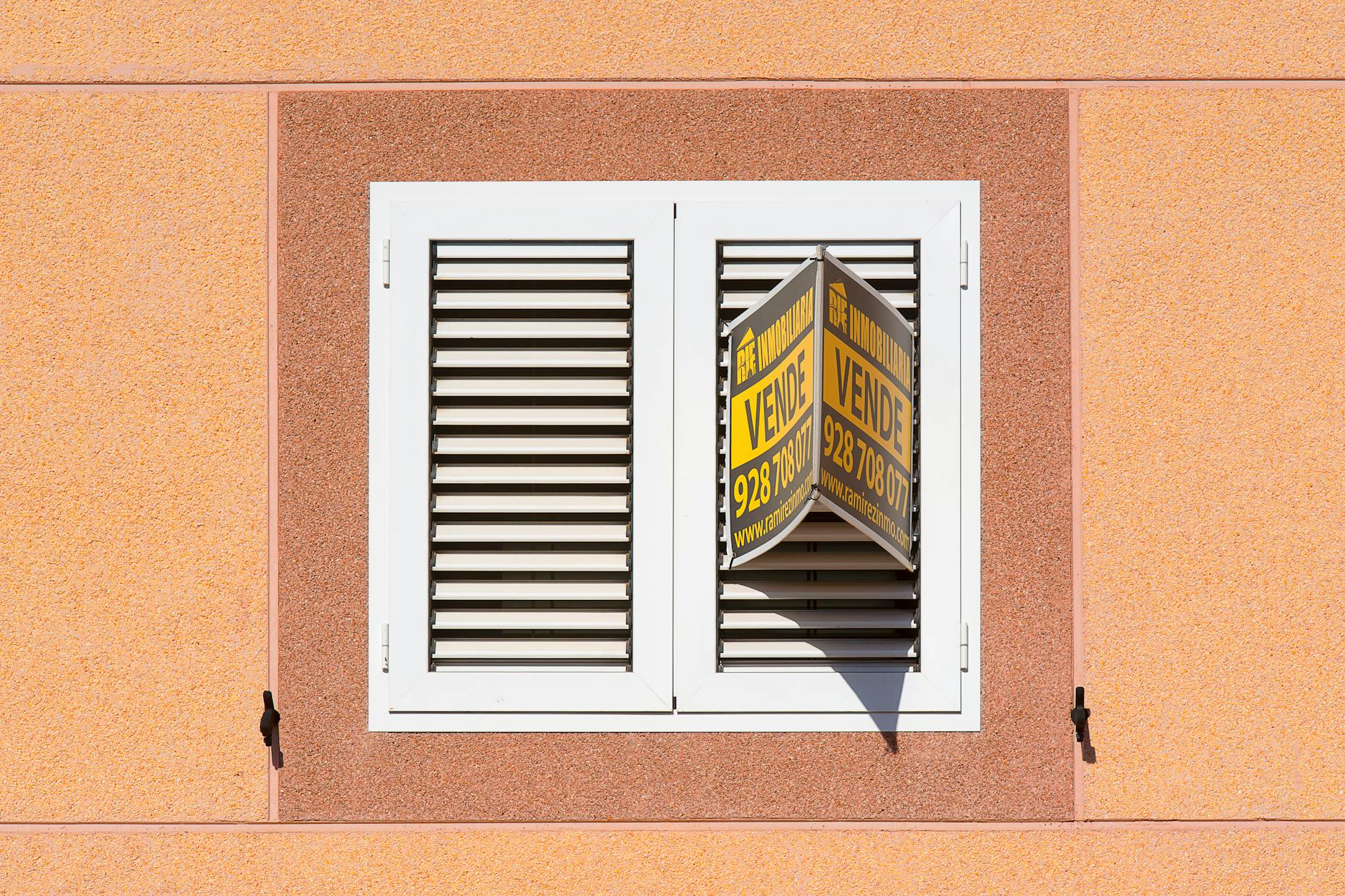 White shuttered window on a terracotta building with a yellow for-sale sign visible between the open shutters