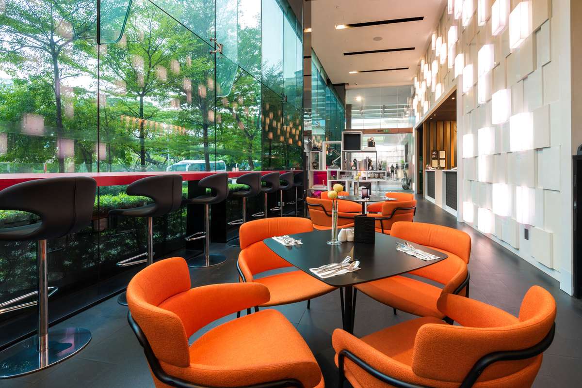 Modern lounge with orange armchairs and a circular table set with drinks in front of a large glass wall overlooking trees.