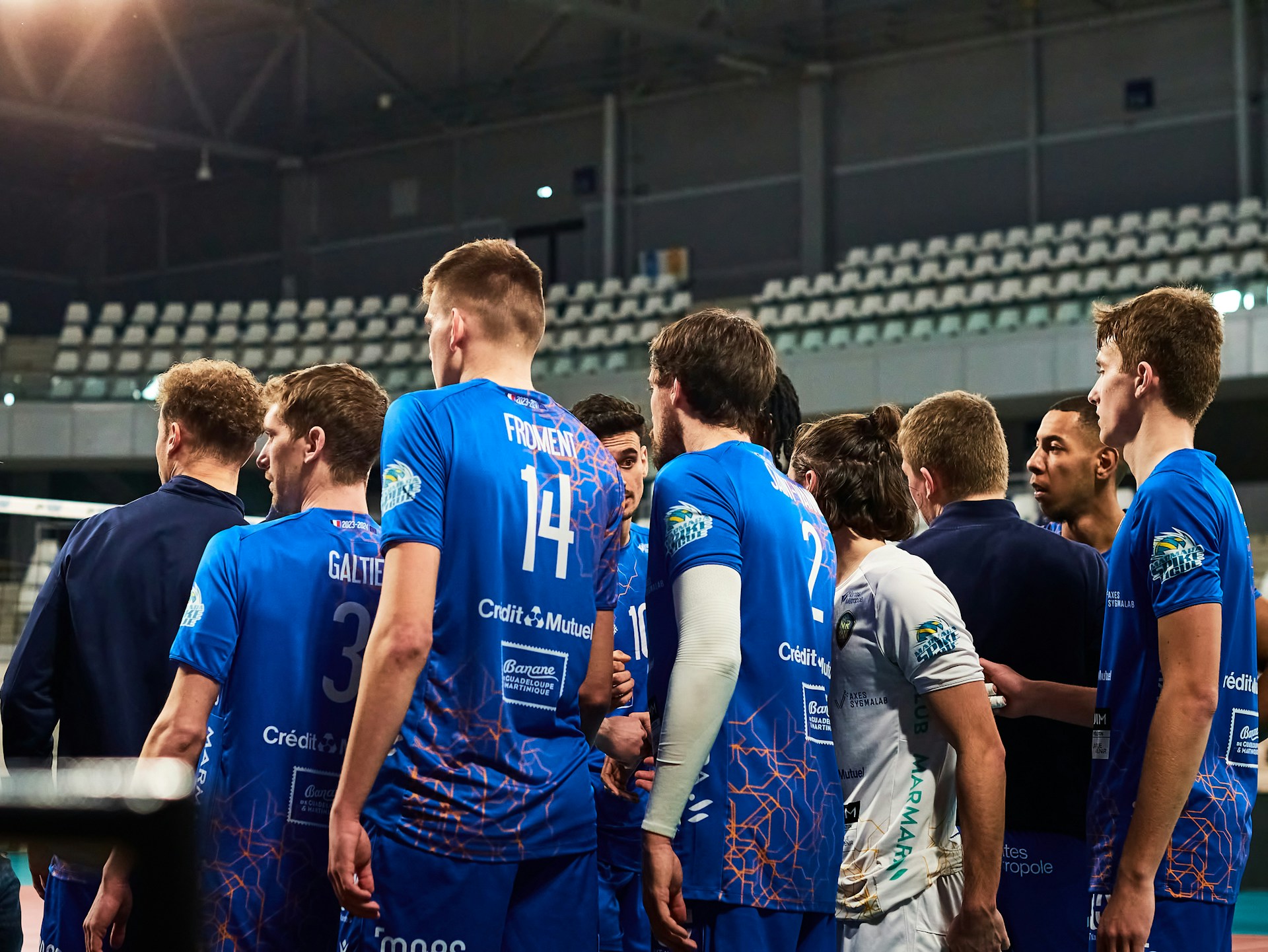 A men's volleyball team wearing blue jerseys gathered in a tight huddle on the court.