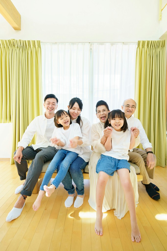 A joyful Asian family, spanning three generations, sits together on a couch in a sunlit living room with green curtains, sharing a moment of laughter and happiness.