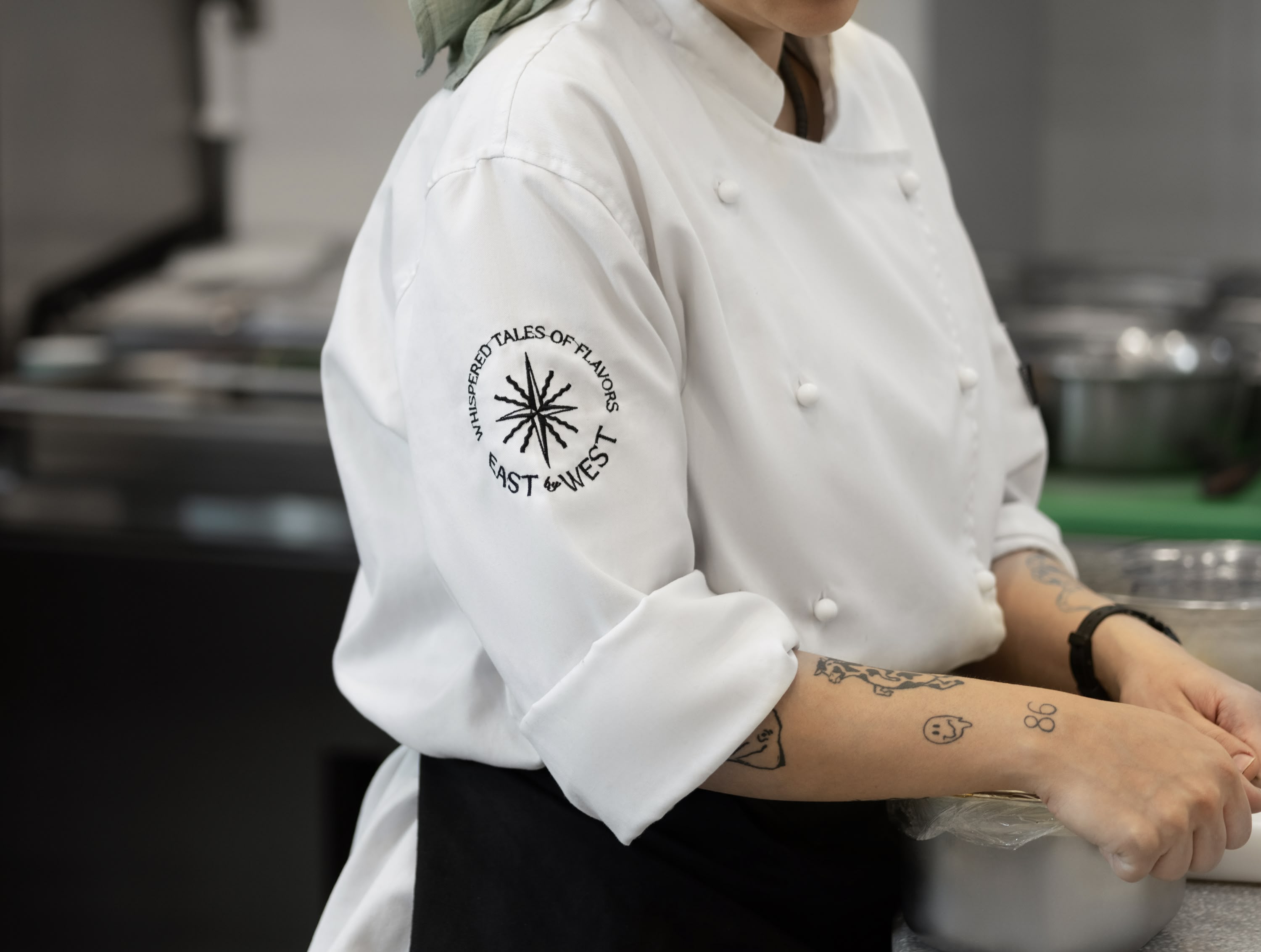 Chef in white uniform with green headscarf wearing East by West "Whispers of Flavors" star-embossed patch on sleeve, preparing food at kitchen counter with pots.