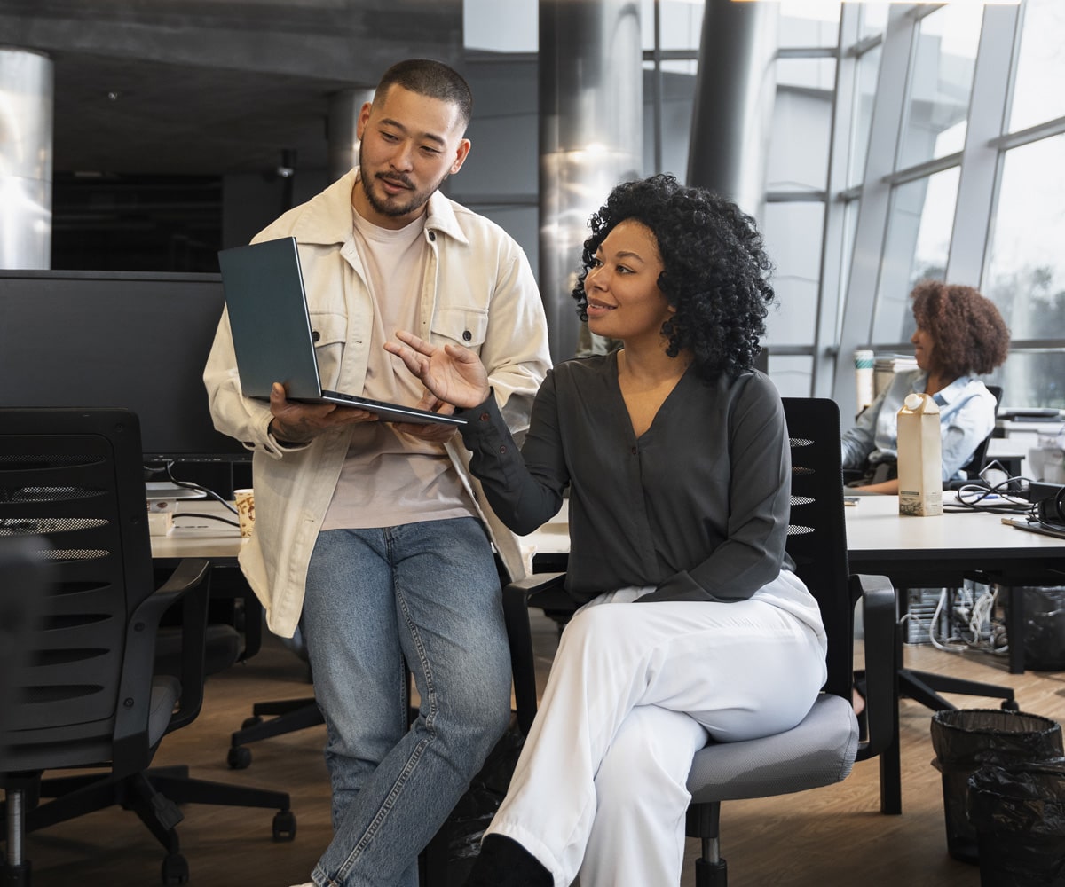 Two colleagues smiling and giving a high-five at a desk in a modern office with a laptop, coffee mug, and plants.