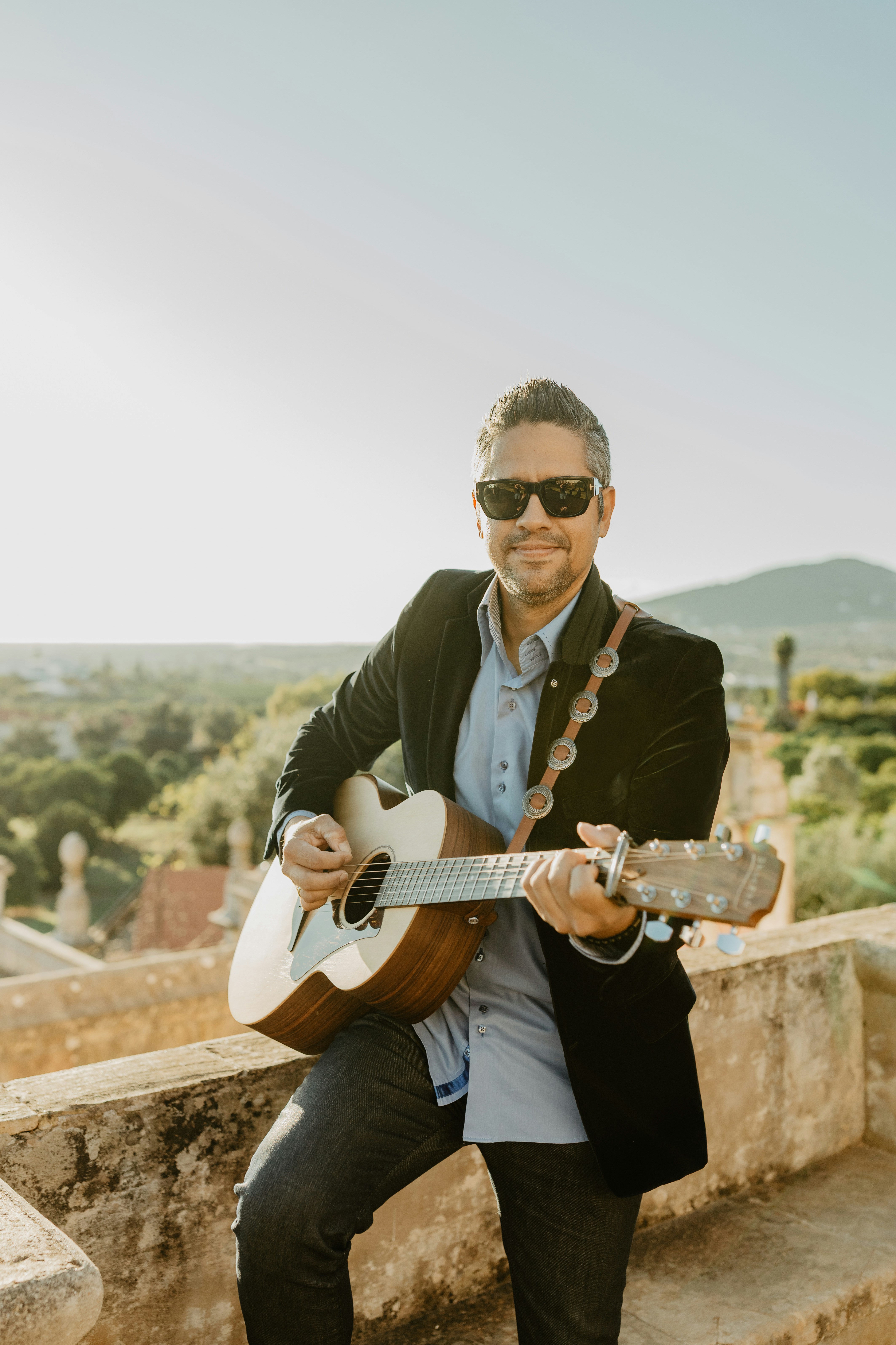 Man playing acoustic guitar outdoors with a scenic background.