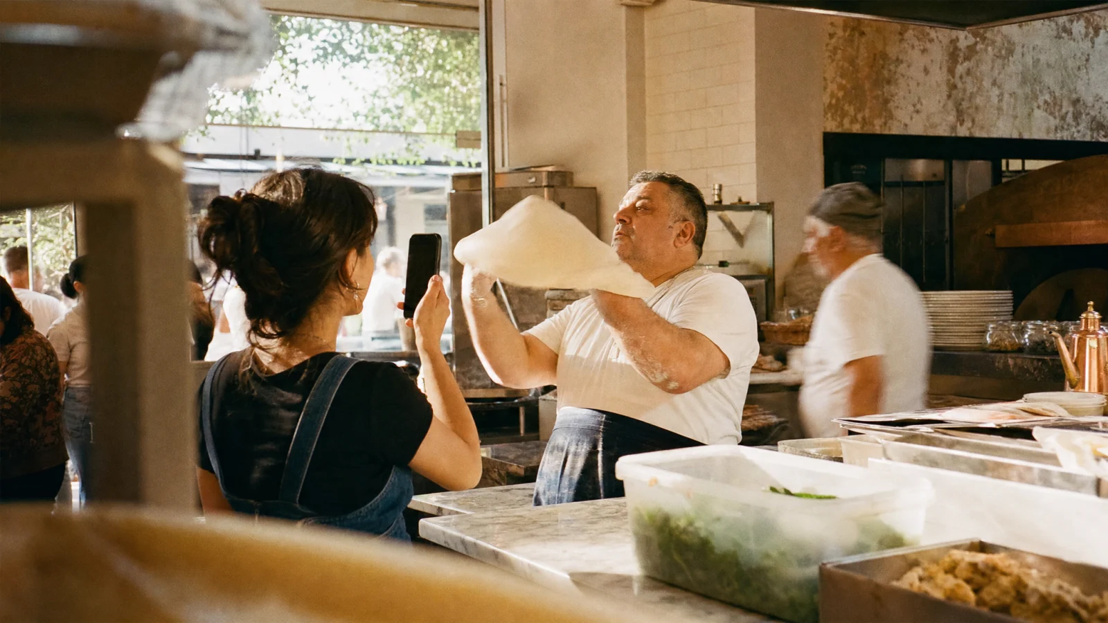 Cameriera riprende con lo smartphone il pizzaiolo che stende l'impasto nella cucina a vista