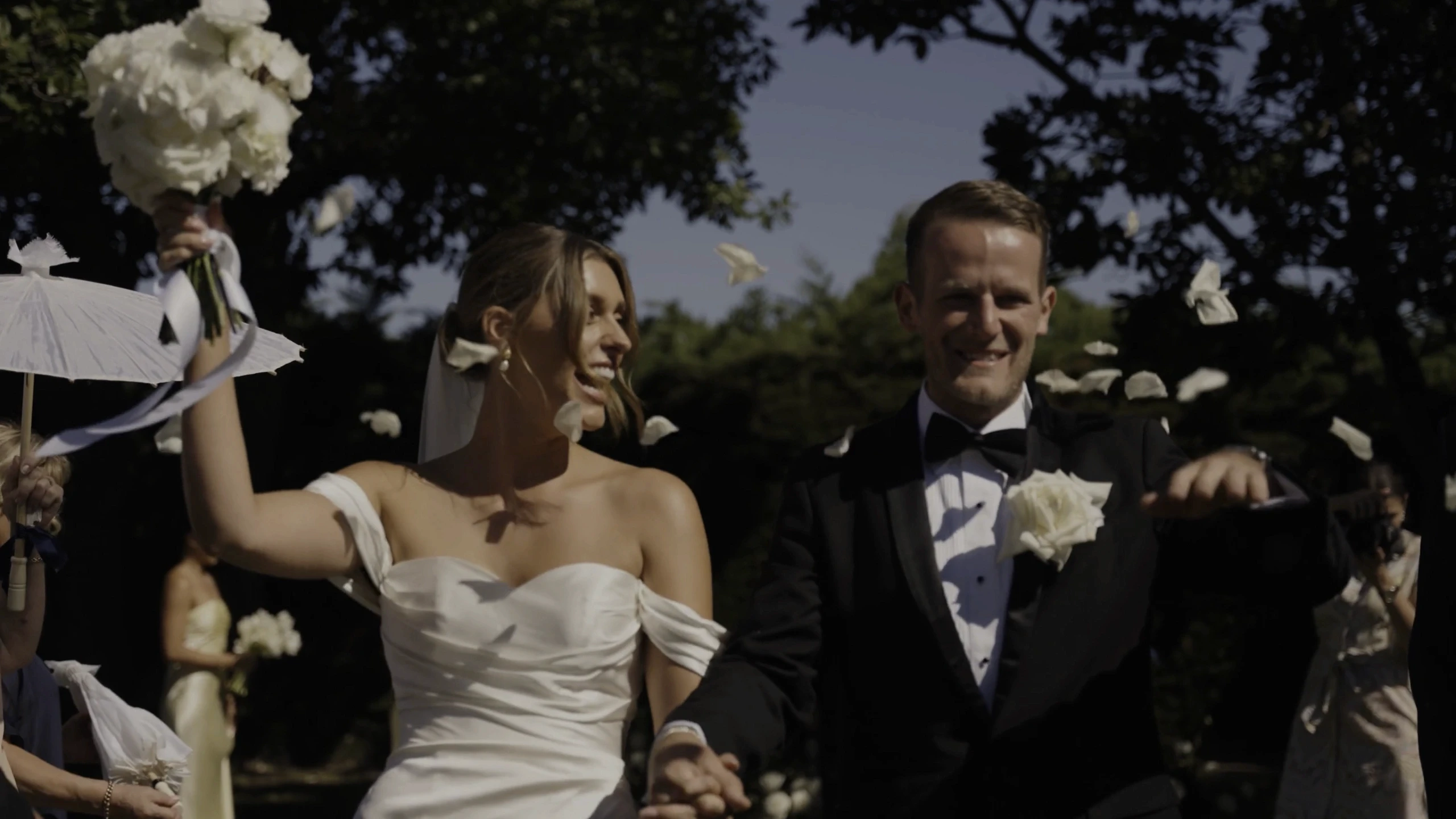 A joyful bride and groom, dressed in an elegant white gown and classic black tuxedo, celebrate their wedding outdoors, surrounded by falling white rose petals and lush greenery.