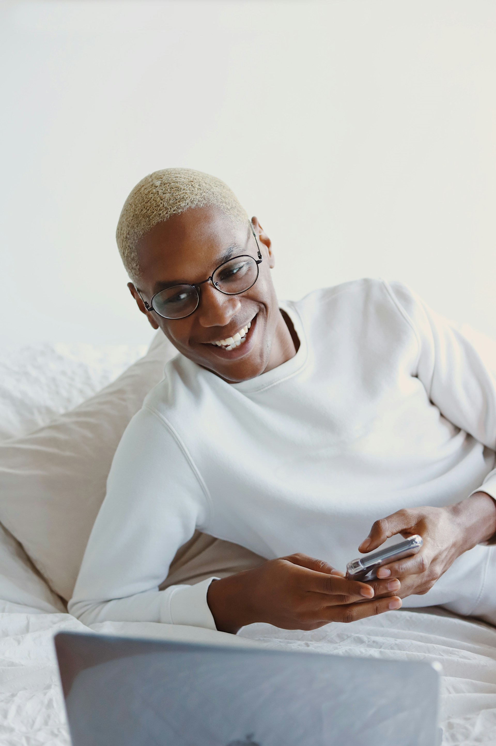 A smiling man with buzzed blonde hair and glasses lies on a bed, looking at a laptop while holding a smartphone. He is wearing a white sweatshirt.