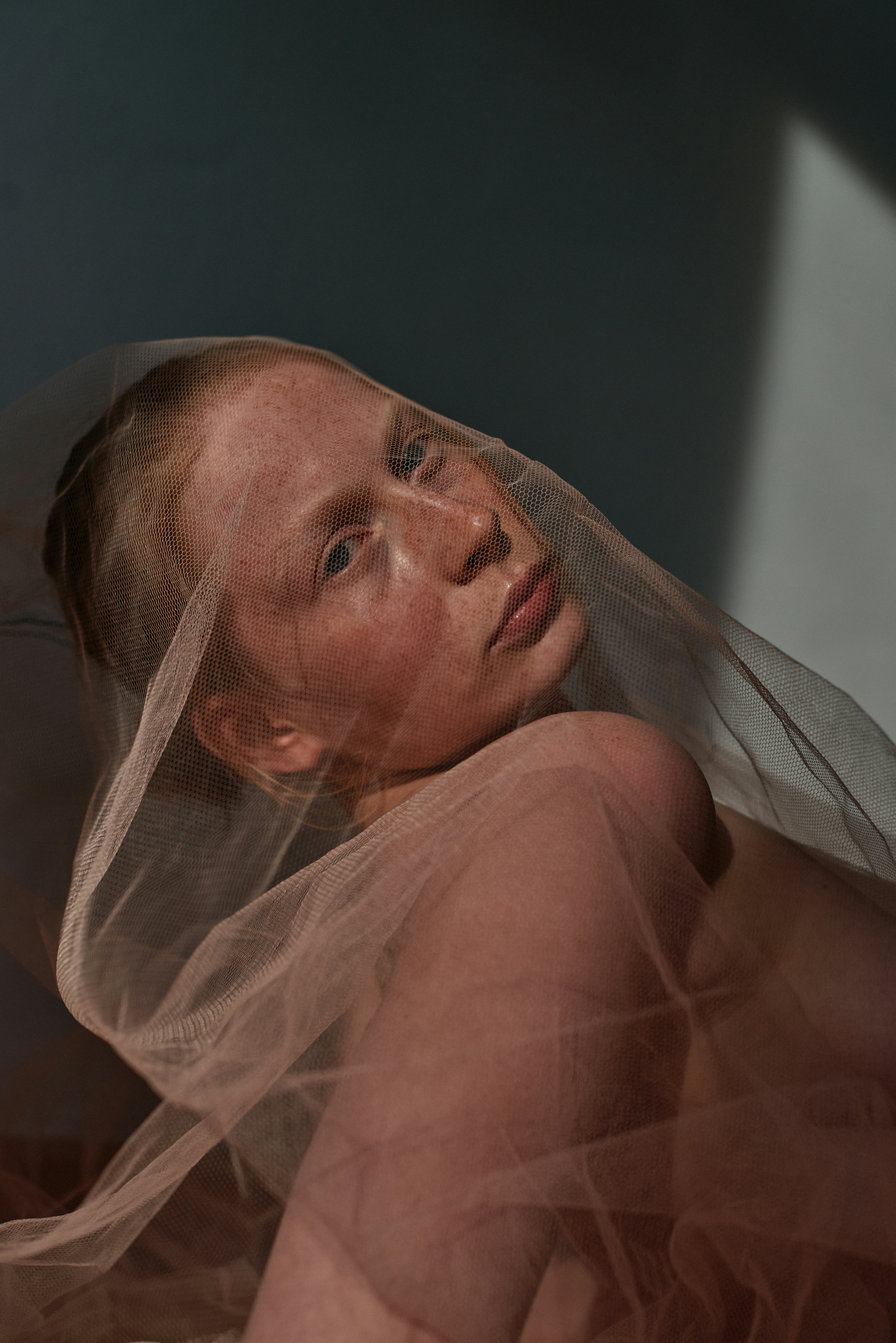 Intimate studio portrait of a freckled model draped in sheer tulle fabric, dramatic directional light against a dark teal wall