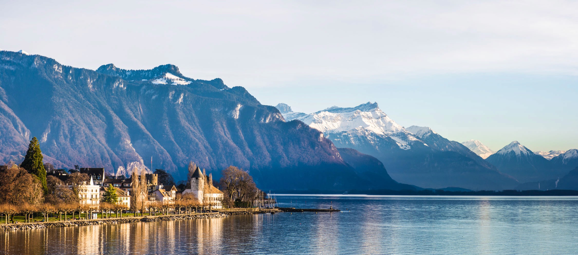 A row of alpine chalets with wooden balconies sits along a lake shore against a backdrop of snow-capped mountains.