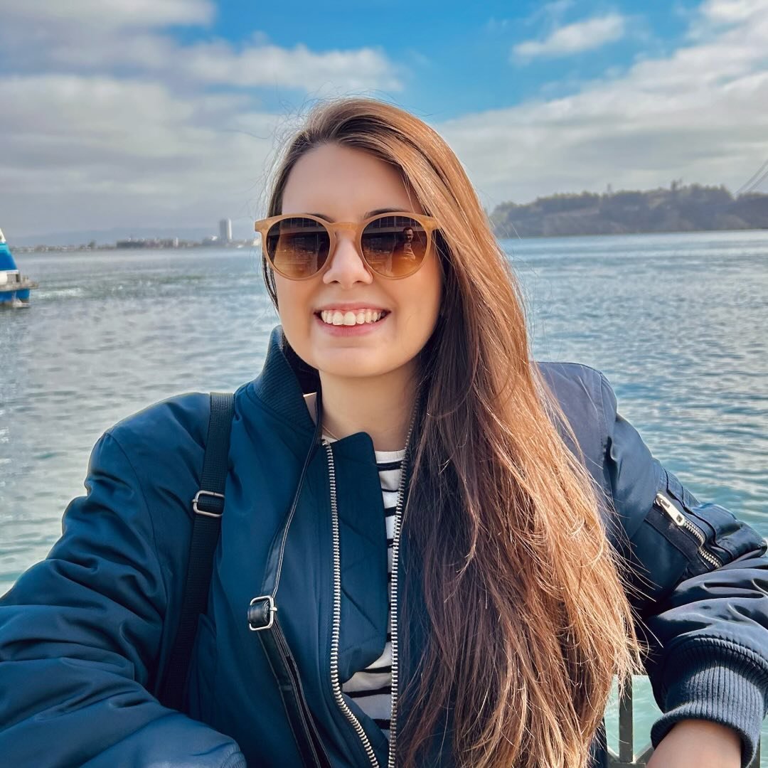 A smiling woman with long hair, wearing sunglasses, poses against a scenic outdoor backdrop with water and clouds.