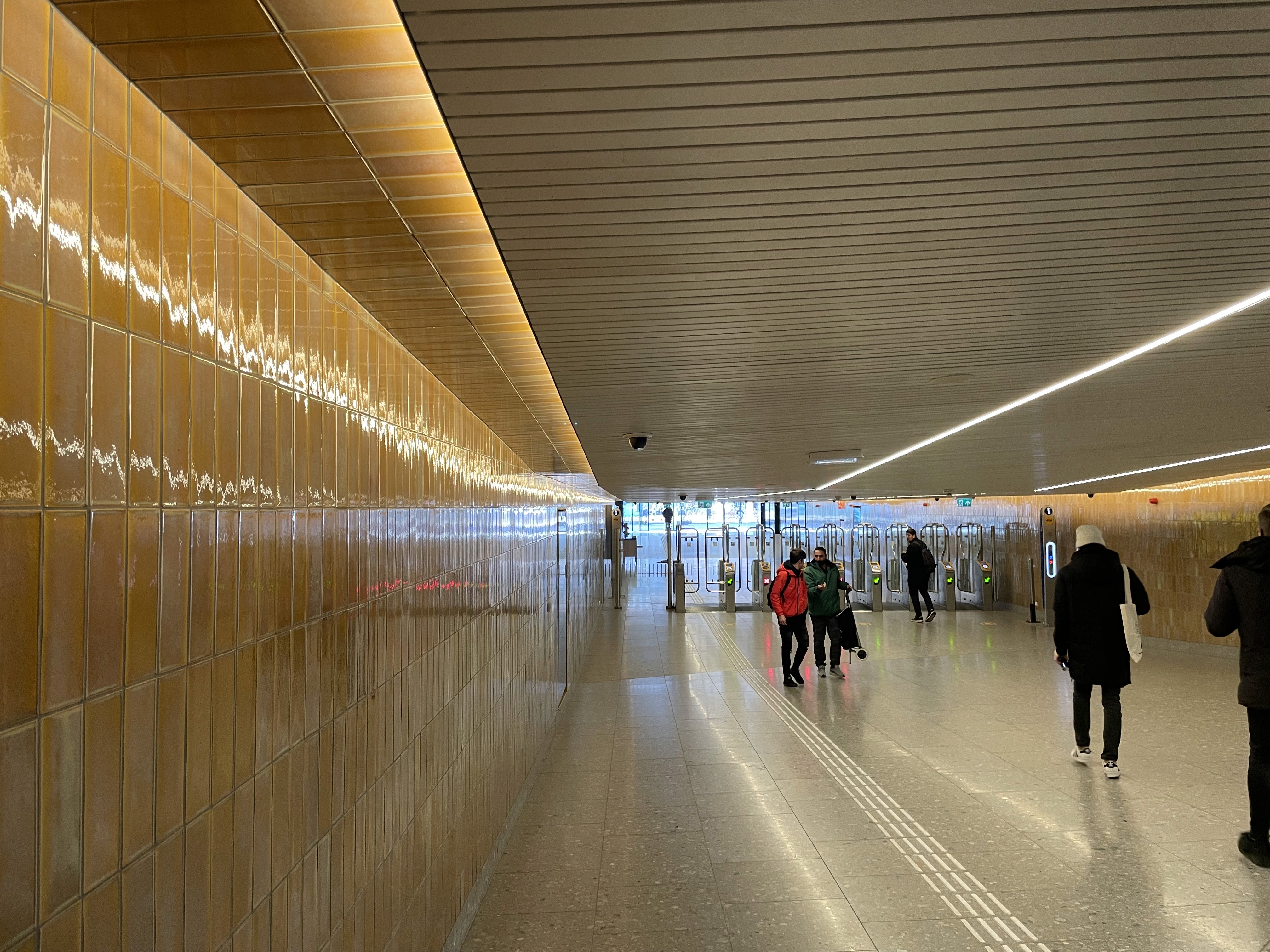 Interieur met gele tegels, mensen en toegangspoortjes in een doorgang.