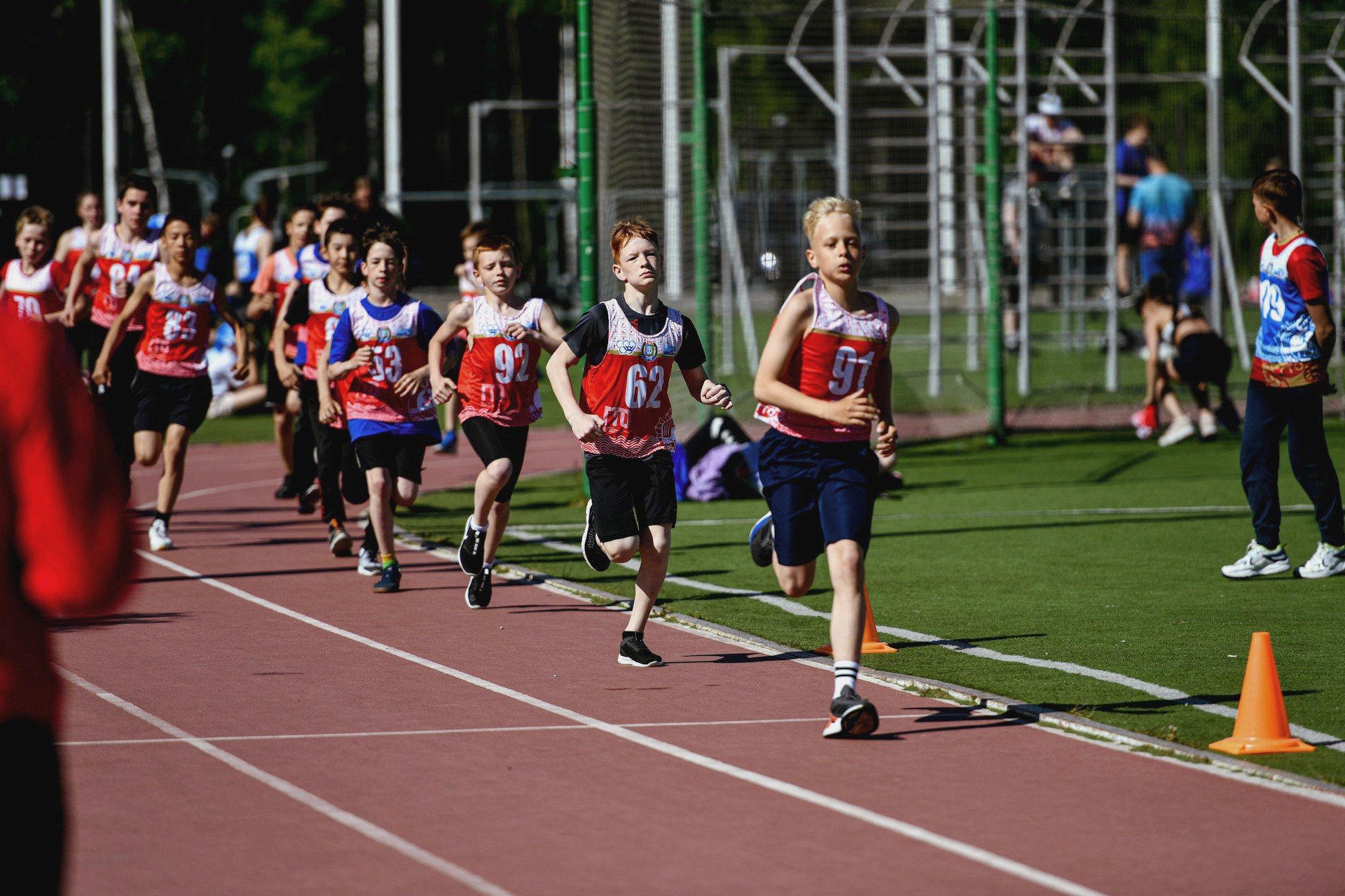 A large group of young boys run a race on an outdoor track, with the focus on a runner in a red and black jersey.