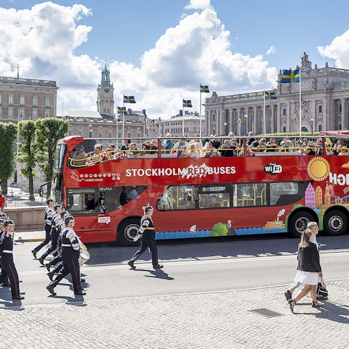 A red double-decker tour bus labeled "Stockholm Red Buses" with tourists on top, passes by a marching band and historic buildings.