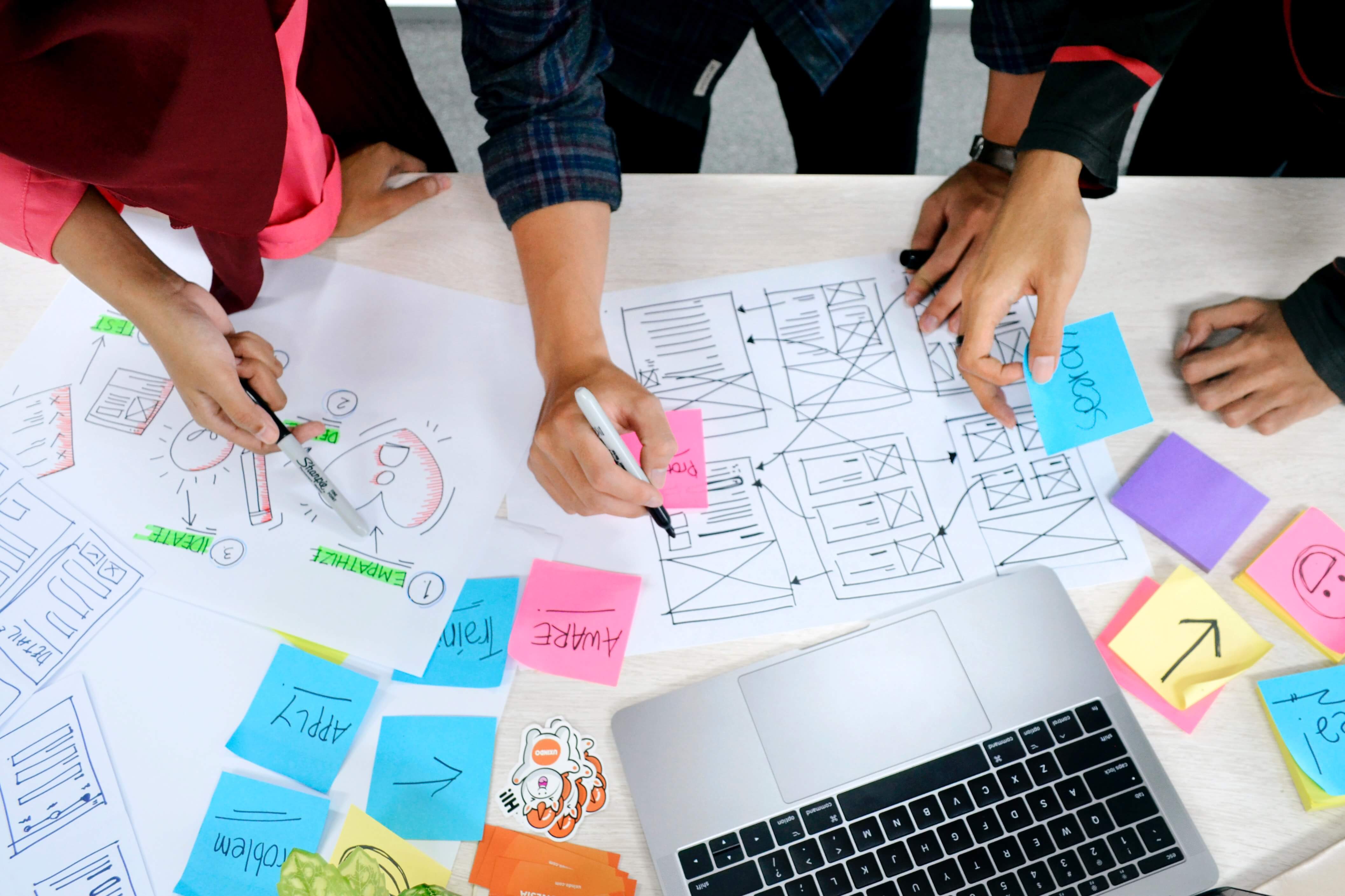 A group of people gathered around a desk with a laptop, sketches and sticky notes on it