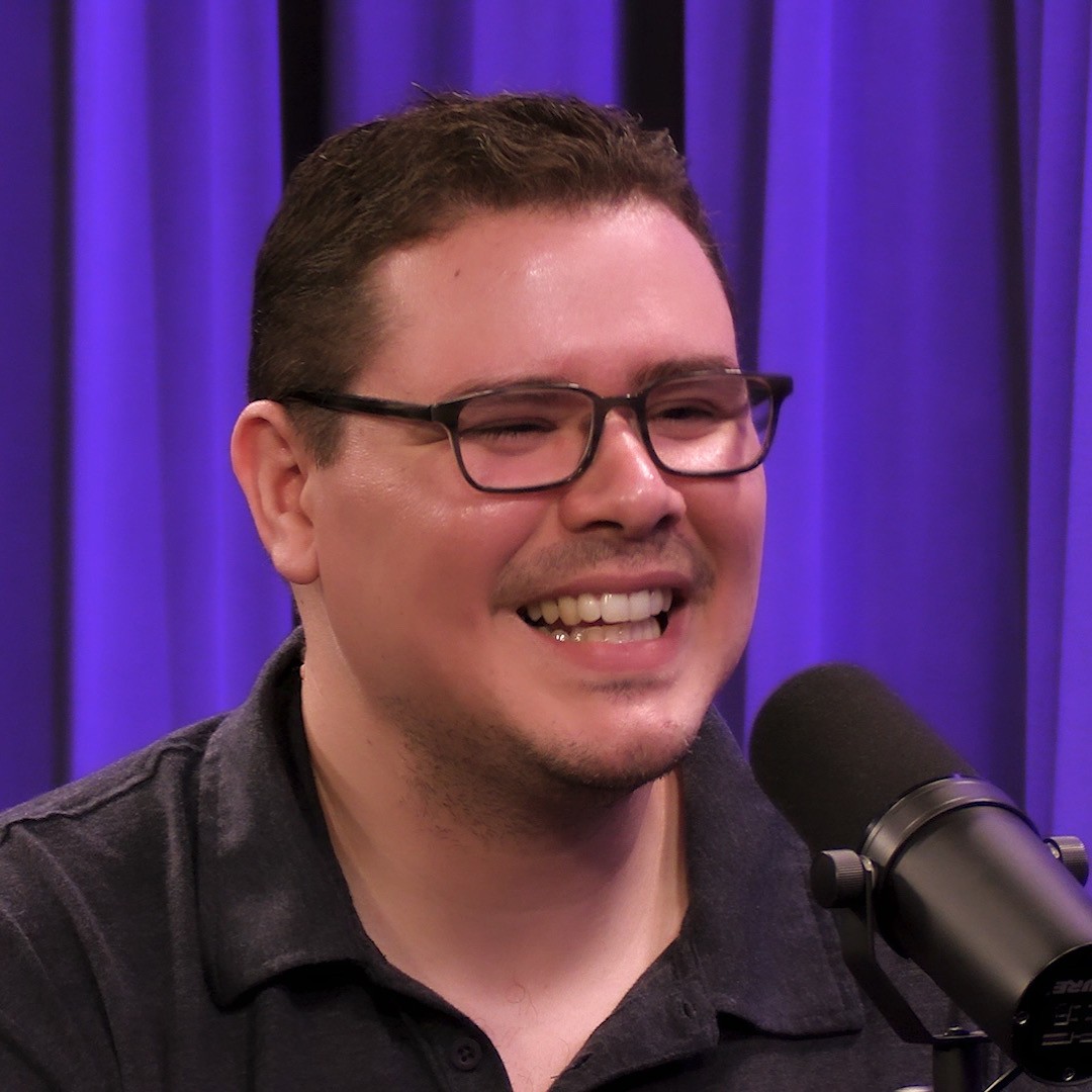 A close-up of a cheerful person with glasses and a dark polo shirt speaking into a professional podcast microphone. The purple curtain background and lighting create a warm, engaging podcast studio vibe.