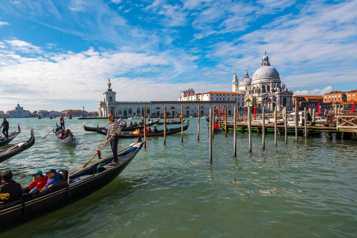 Several gondolas and a large white dome building with a clock tower sit along a canal under a blue sky with clouds.