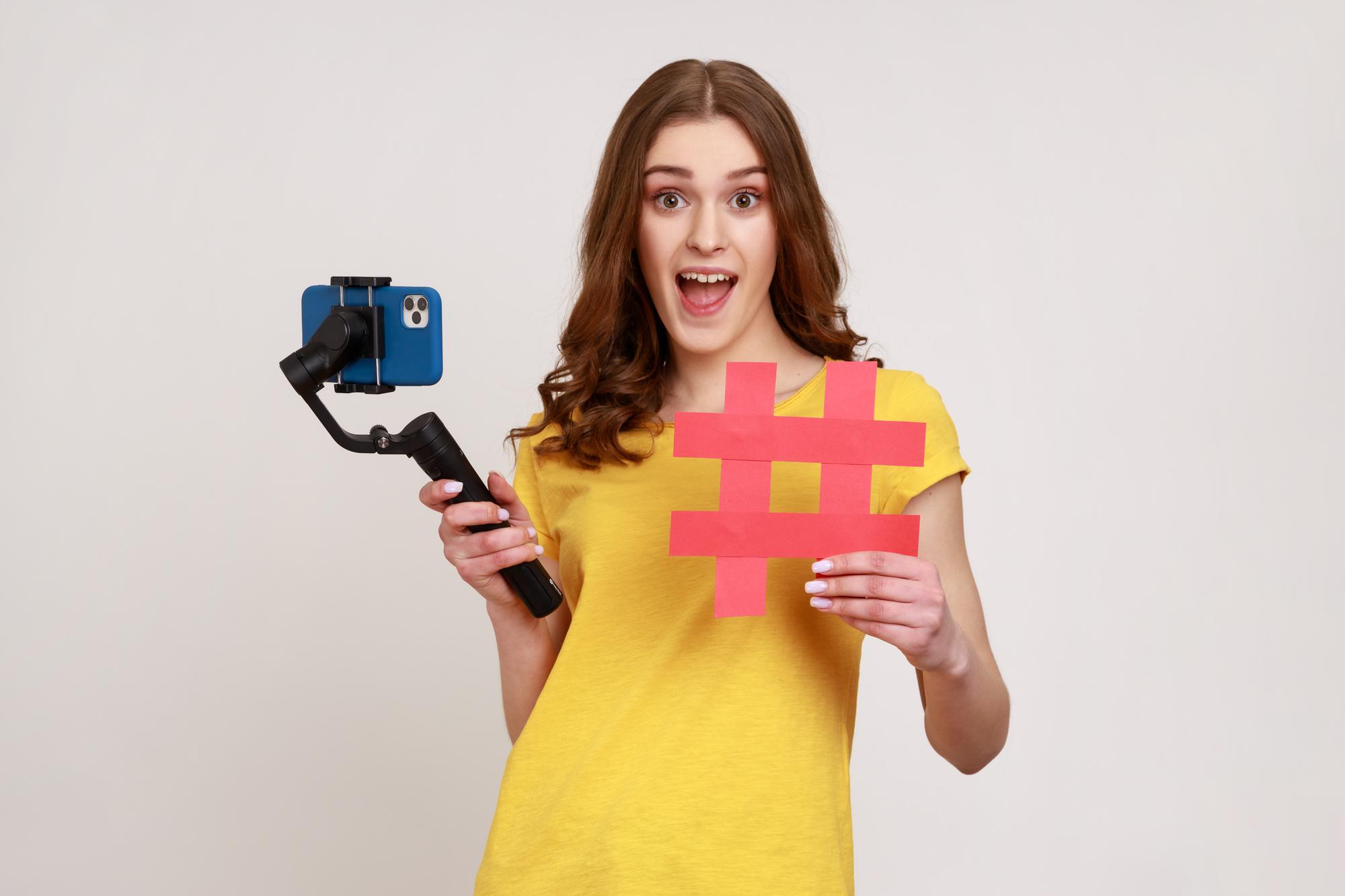 A woman smiles while holding a selfie stick in one hand and a hashtag sign in the other, capturing a fun moment.