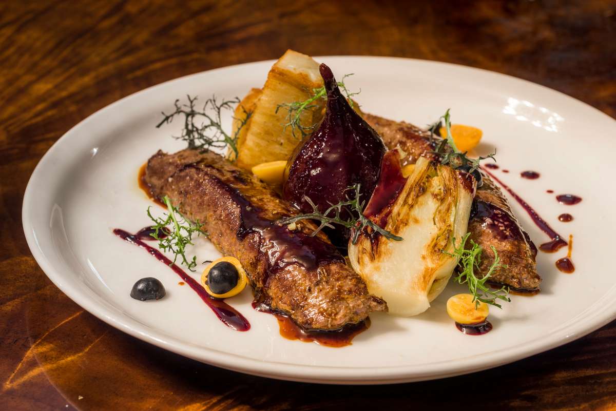 A plated gourmet dish featuring roasted meat, grilled vegetables, and a dollop of orange puree on a white plate.