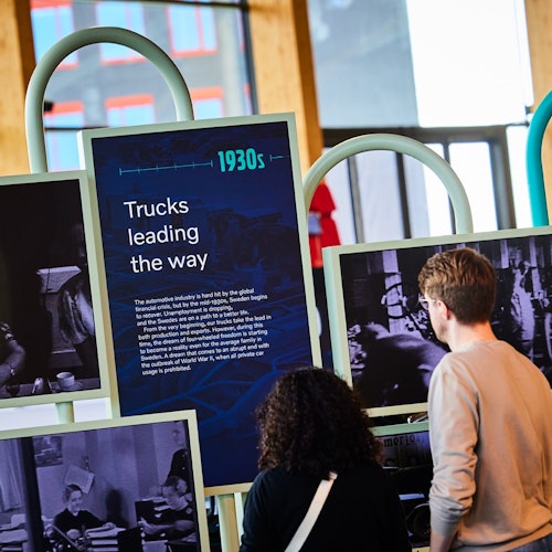 Två personer betraktar en utställning med historiska fotografier och en skylt med texten ”1930-tal: Lastbilar visar vägen” i en modern inomhusmiljö.