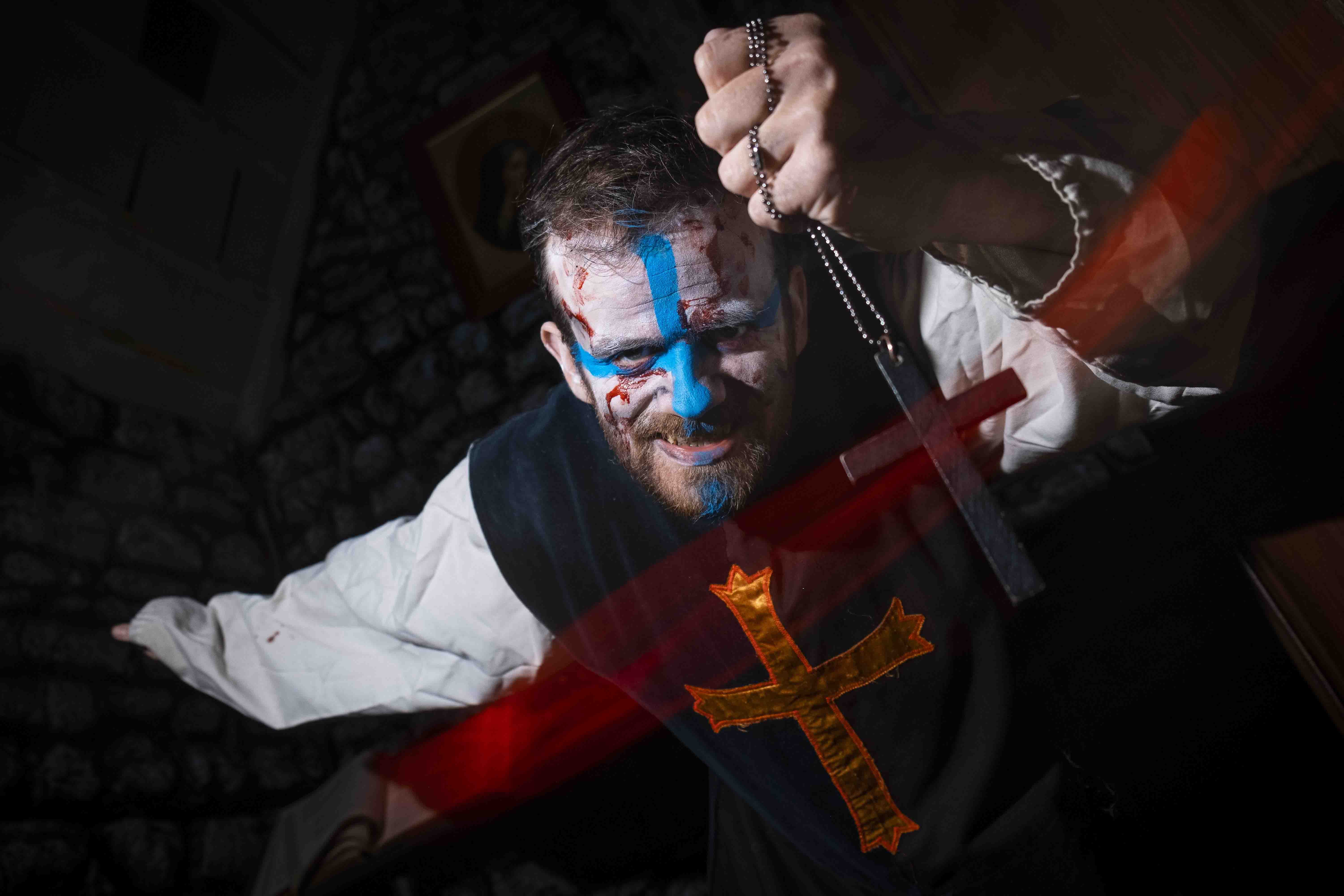 A person in medieval-style clothing and face paint stands against a stone wall, holding a rosary with an intense expression, illuminated by dynamic red and blue lighting.