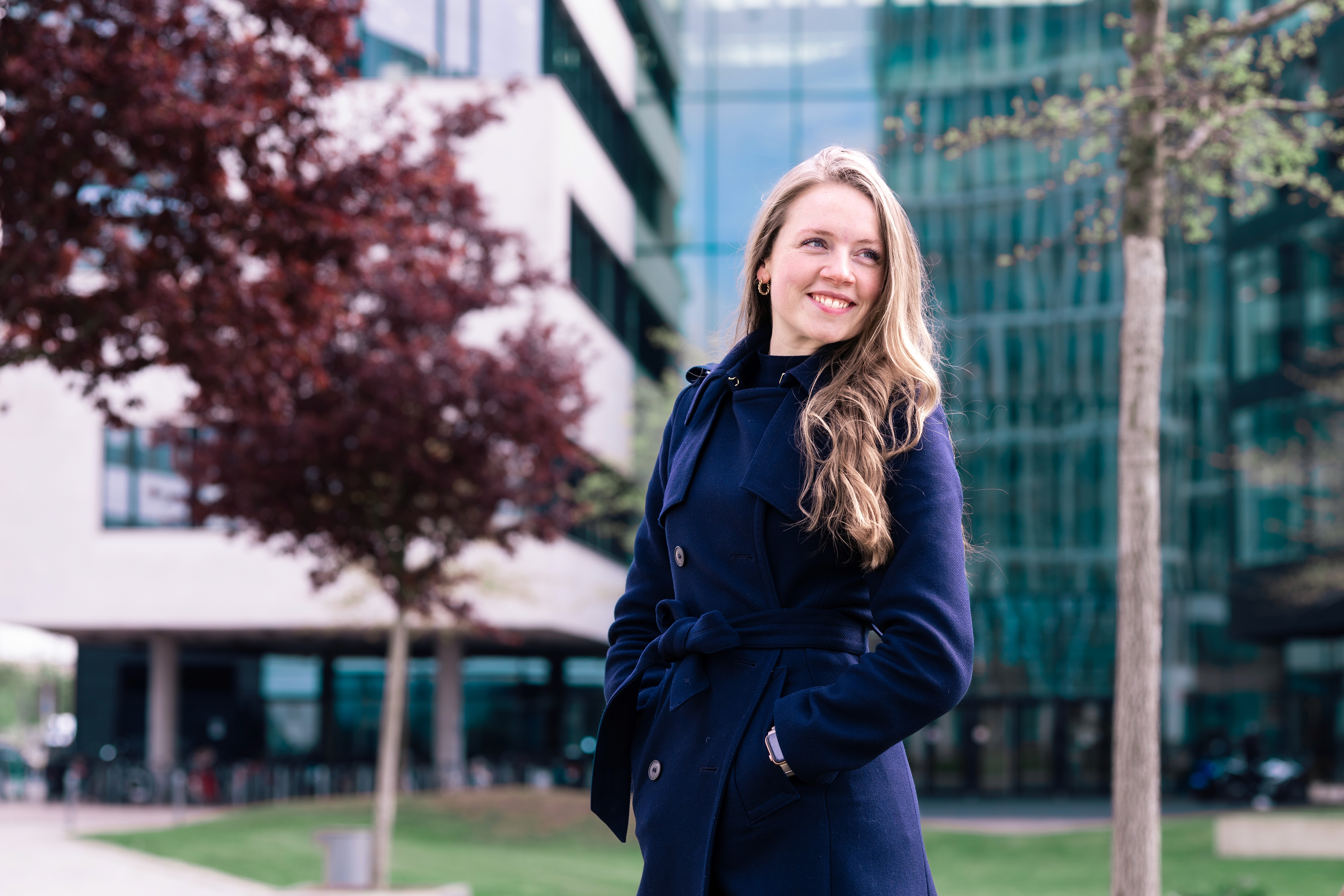 A DevX team member smiling outdoors in a navy coat beside a modern glass office building.