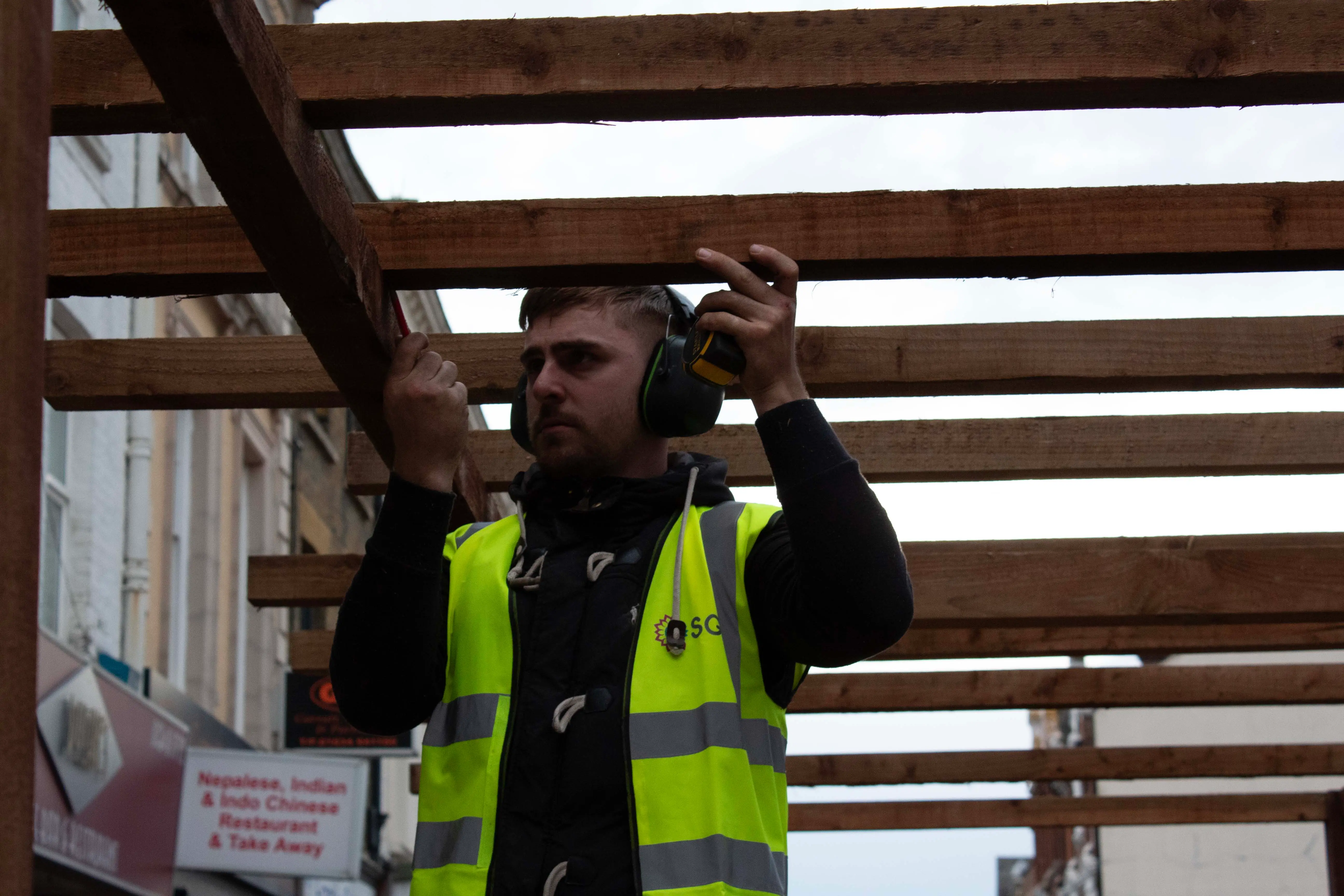 A construction worker in a neon yellow vest raises a tool while standing under a wooden framework.