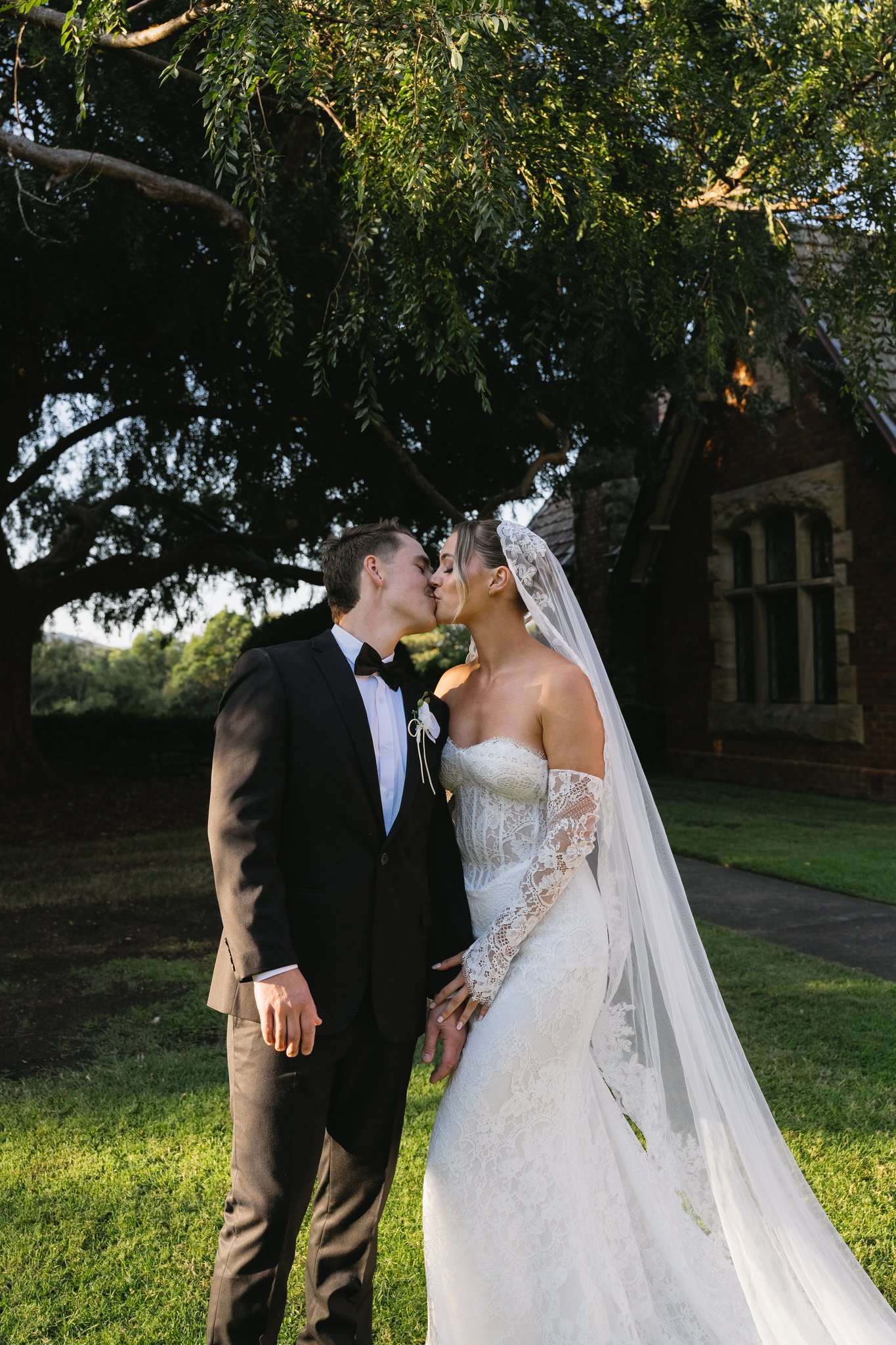 Bride and groom kiss beneath a large tree on green lawn beside a historic brick building with leadlight windows.