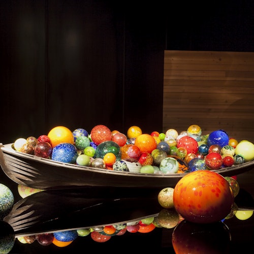 A bowl filled with colorful, variously sized marbles on a glossy black surface, with additional marbles scattered around.